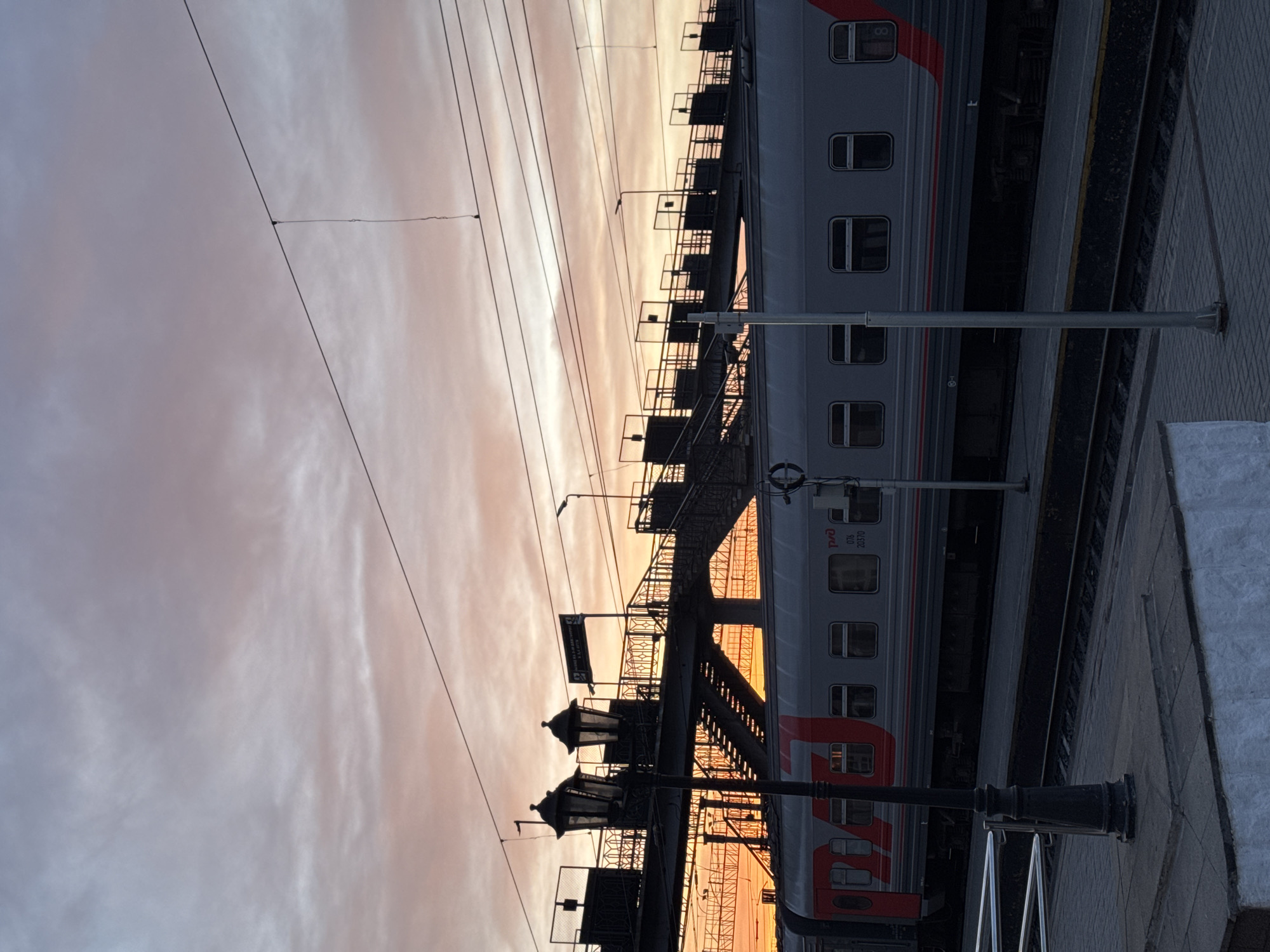 The image depicts a passenger train at a station in Izhevsk, Russia, viewed at dusk or dawn. The sky displays a range of colors from light gray to orange. Overhead power lines are visible above the train and a pedestrian bridge crosses over the tracks. The train is silver and red with rectangular windows. The station platform is paved with concrete blocks. Streetlights are positioned along the platform and bridge.