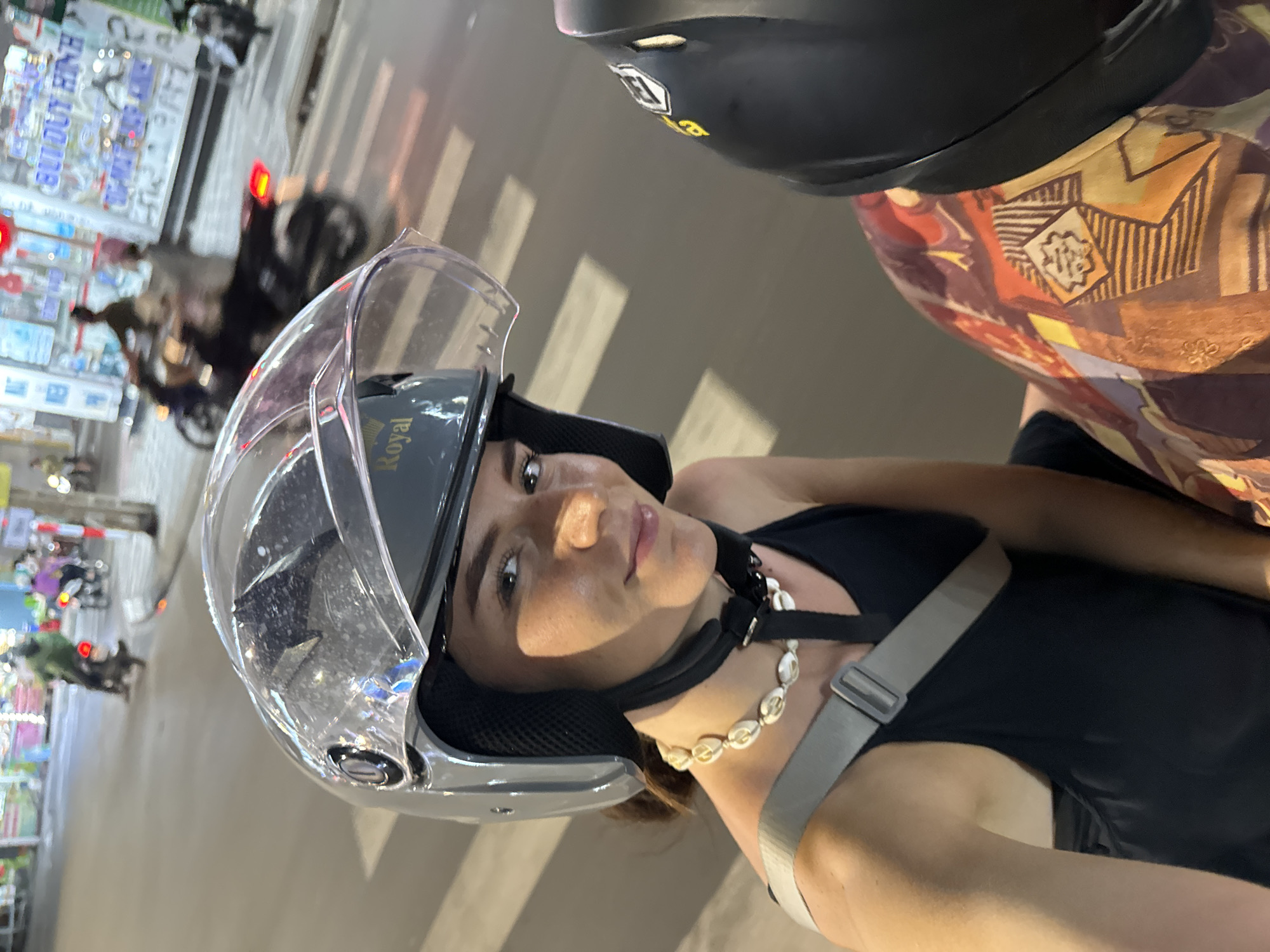The image features a woman wearing a grey open-face helmet with a clear visor, looking directly at the camera. She wears a black halter-neck top and a white seashell necklace. The helmet has the word "Royal" inscribed in gold lettering on its side. Behind and above her, the head and shoulder of another person are visible, wearing a black helmet with a yellow logo and a patterned, multi-colored shirt with abstract designs. This indicates they are likely riding a motorcycle or scooter together.
The setting is an urban street scene in Phan Thiết, Vietnam, captured at night or dusk, suggested by the artificial lighting and blurred vehicle lights in the background. The street appears busy, with other motorcycles and blurred figures moving about. Along the street, storefronts and buildings are visible. One prominent sign in the background displays partially legible Vietnamese text, "BUUUYT HINH," likely part of a business name. Faint white street markings, resembling a zebra crossing, are visible on the road surface. The woman maintains a neutral expression while engaging with the camera. The perspective suggests a selfie or a photo taken by the person behind her on the same vehicle.