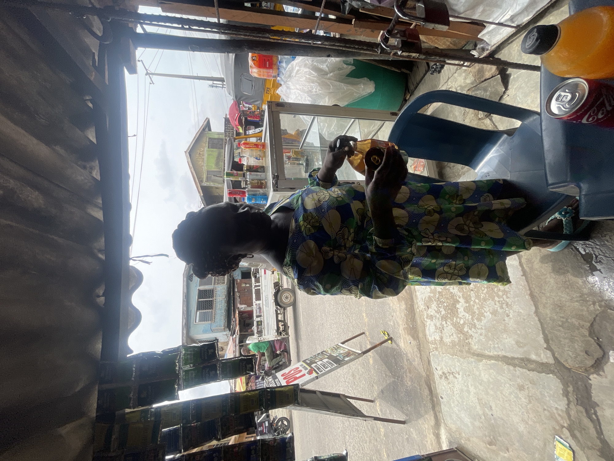 In Akowonjo, Nigeria, a woman stands inside a small storefront, holding a bottle of brown liquid. She is wearing a colorful, floral-patterned dress and her hair is braided. In front of her is a clear glass display case filled with bottles and other items. To her right, a blue plastic chair sits empty, with a can of Coca-Cola and a bottle of orange-colored liquid on its seat. Behind her, through the open doorway, a street scene unfolds. A man rides a tricycle in the street, and various buildings and vehicles are visible. The storefront is covered by a corrugated metal roof, and items hang from the ceiling. The ground outside is paved and appears wet in places. The overall impression is of a bustling local market or shop.