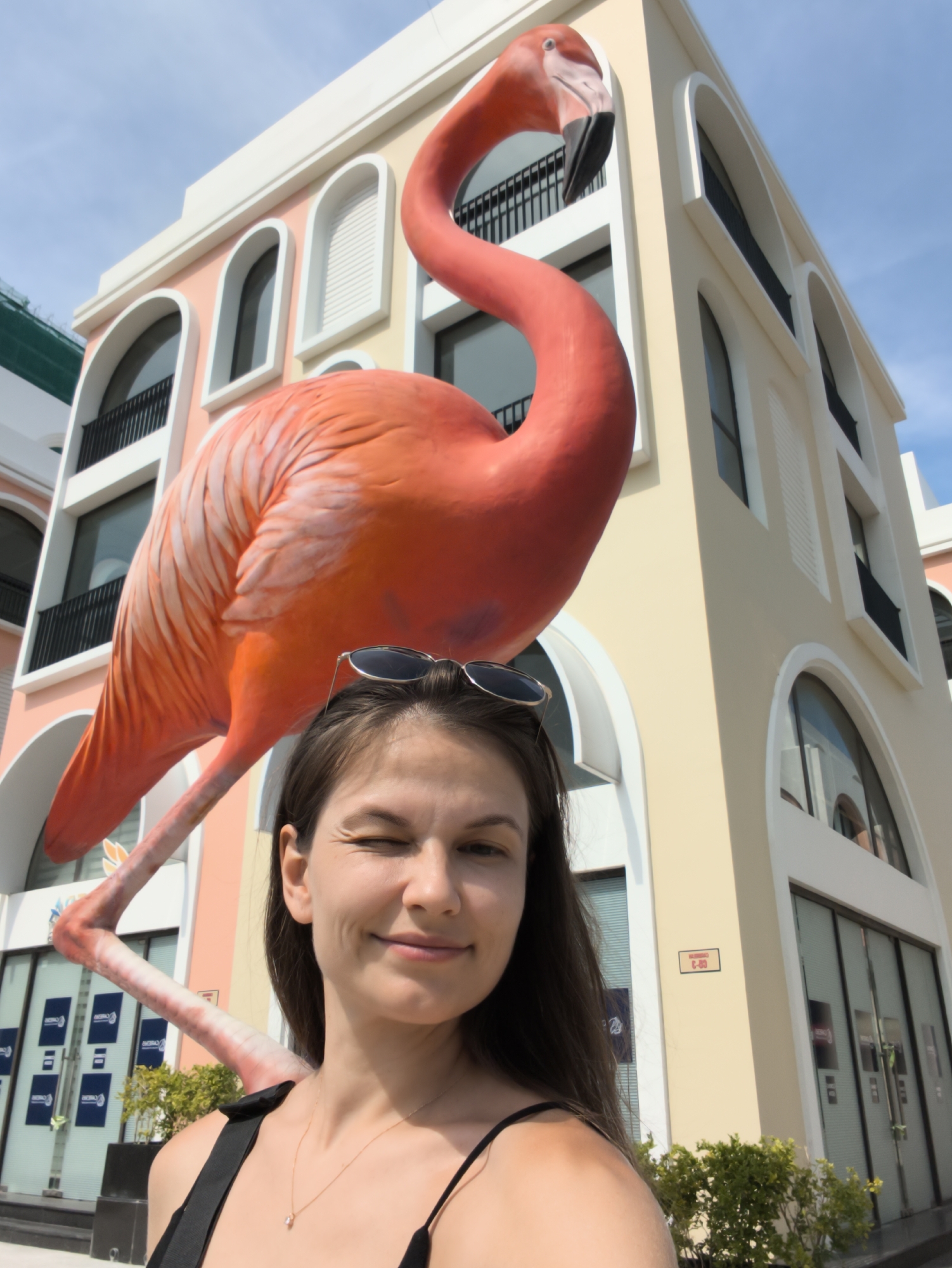 A medium shot captures a young woman in the foreground, winking at the camera, with sunglasses perched on her head. Behind her, a large, vibrant orange flamingo statue is prominently displayed. The flamingo appears to be part of a decorative installation, with its long neck curving over the woman's head. In the background, a multi-story building with a pastel pink and yellow facade stands against a clear blue sky. The building features arched windows and balconies, suggesting an architectural style that might be found in a resort or entertainment district. The overall scene conveys a sense of fun and tourism.