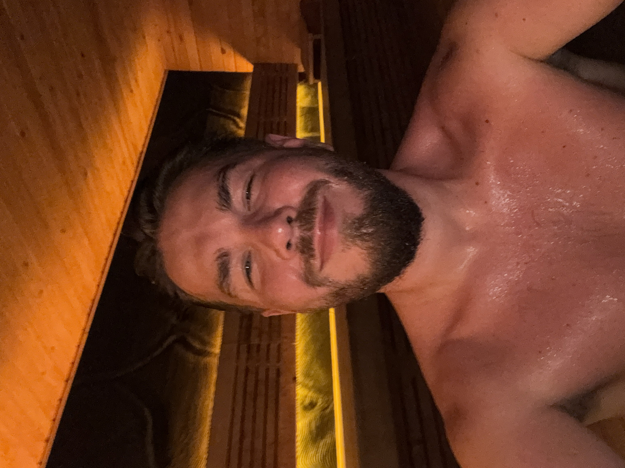 A medium shot shows a man with a beard in a sauna. The man is lying down, facing the camera with a slight smile. His skin is red and appears to be covered in sweat. The sauna is made of wood with wooden benches and a light strip in the background. The location is unknown and there is no indication of it being in Mengwi, Indonesia.