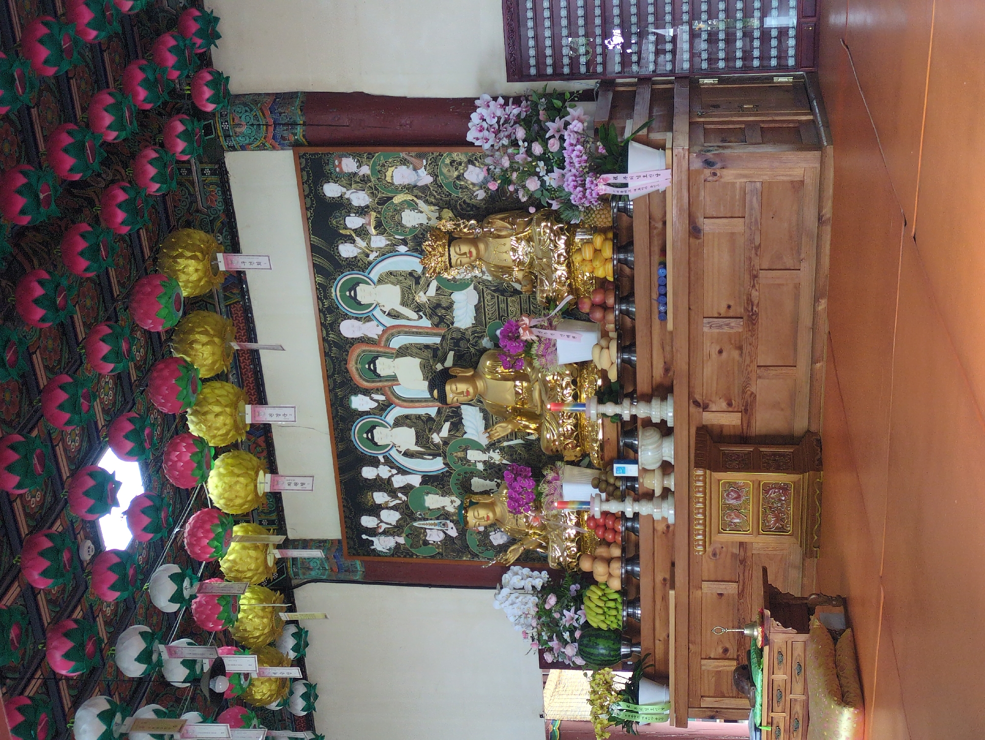 The image depicts an interior Buddhist shrine in Gongju-si, South Korea. Three gold Buddha statues are displayed on a wooden stand, each accompanied by offerings of fruit and flowers. A painted backdrop featuring additional Buddha figures is behind the statues. Hanging from the ceiling are numerous lotus-shaped lanterns in pink, green, yellow, and white. A small wooden cabinet and a cushion can be seen on the right side of the image.
