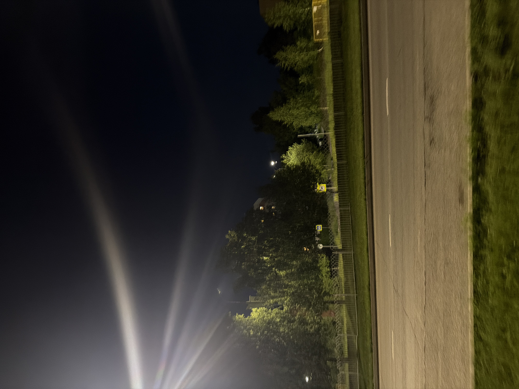 The image captures a nighttime scene on a street in Naberezhnye Chelny, Russia. A bright light source, possibly a streetlamp, illuminates the left side of the frame with strong beams. The street, with visible white lane markings, stretches across the lower portion of the image. Adjacent to the street is a strip of grass, followed by a fence and a line of trees. Beyond the trees, a building with illuminated windows is visible. The sky above is dark, with faint, streaky clouds. Two yellow road signs are present; one is circular with a "40" inside, and the other is a yellow square with a black symbol.
