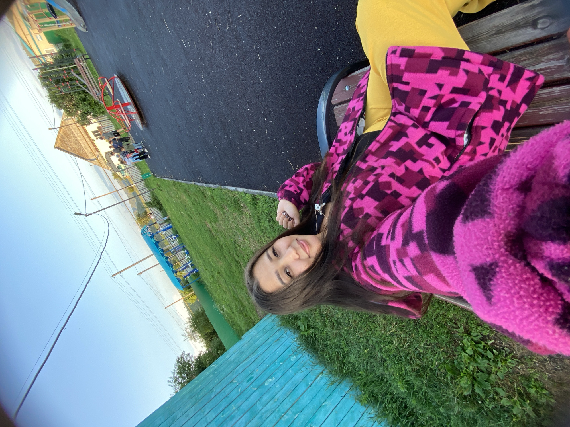 The image shows a young woman sitting on a park bench, looking at the camera. She is wearing a bright pink and purple patterned jacket and yellow pants. The bench is made of wood and metal and is located on a paved area. Behind the woman, there is a playground with a merry-go-round and other equipment. There are several children playing in the background. In the distance, there are houses and trees, suggesting a rural or suburban setting. The sky is clear and blue. The location is identified as Maloarslangulovo, Russia.