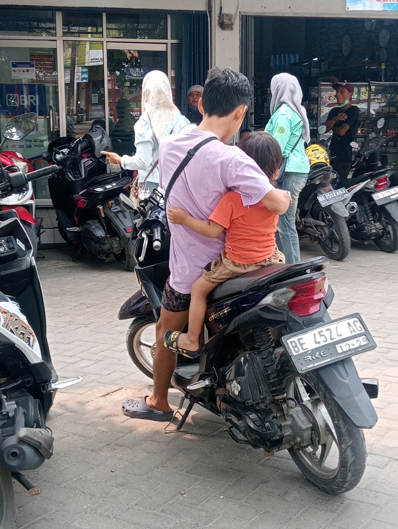 In Bandar Lampung, Indonesia, a man and a child are on a motorcycle. The man, wearing a purple shirt and brown patterned shorts, is embracing the child, who is wearing an orange shirt and beige shorts. They are positioned on a dark-colored motorcycle in the foreground. Behind them, several other motorcycles are parked. In the background, a building with large windows and glass doors appears to be a commercial establishment, possibly a shop. The ground is paved with grey interlocking bricks. The scene is illuminated by natural daylight, casting shadows on the ground. Several other individuals, some wearing headscarves, are visible in the background, suggesting a public area. The motorcycles have license plates. One plate reads "BE 4524 AG".