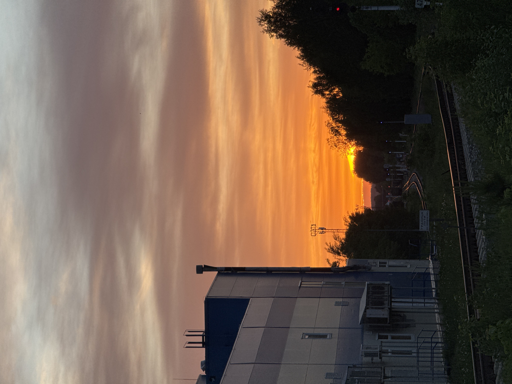 The image shows a sunset in what appears to be an industrial area in the city of Chaikovsky, Russia. In the foreground is a multi-story building with a blue and white striped design. Beside the building is railway tracks. A forested area runs alongside the railroad. The sky is a mix of orange, pink, and gray, indicating either a sunrise or sunset. The illumination is generally dim, consistent with twilight conditions.
