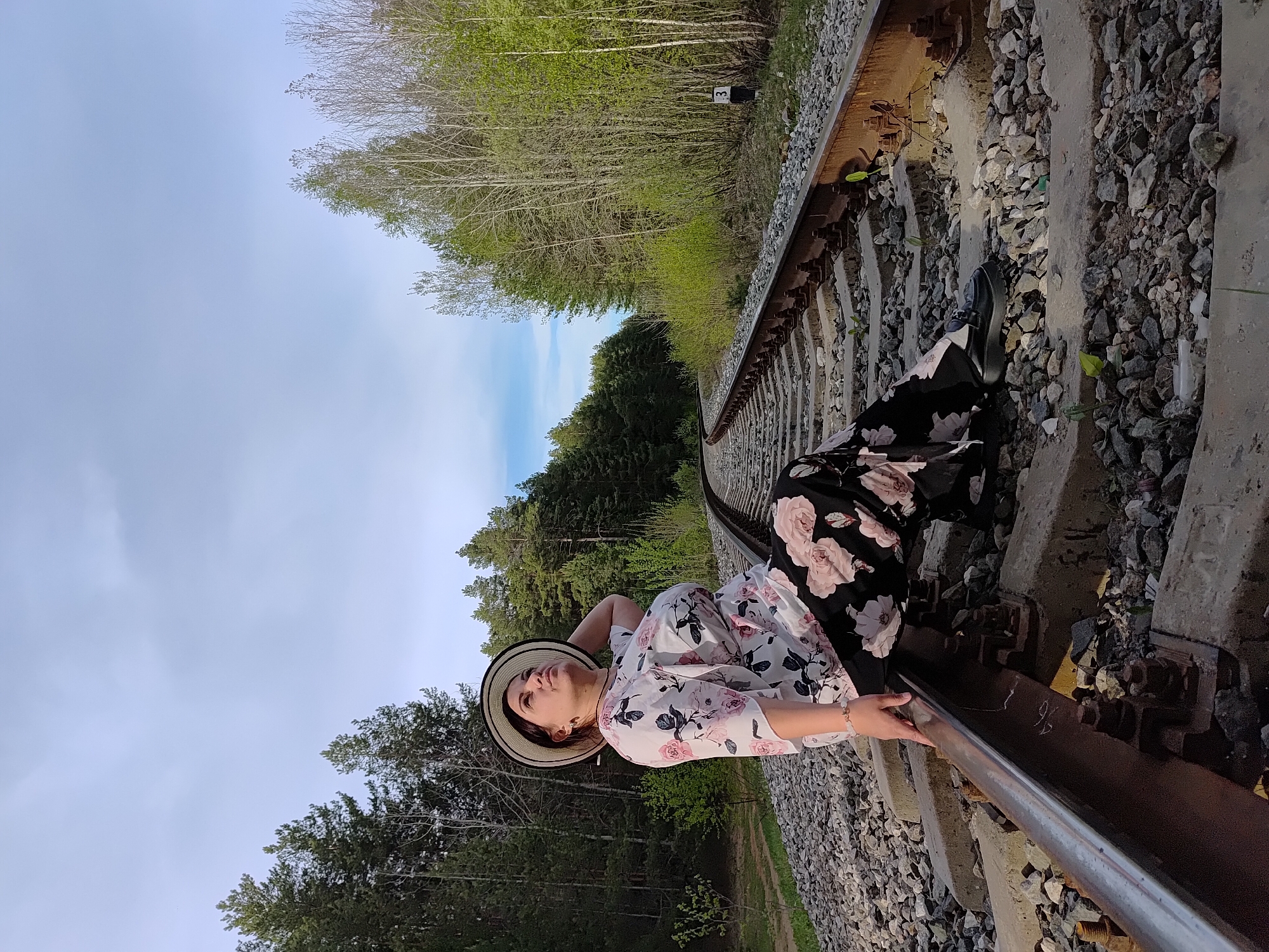 A woman is lying on railroad tracks in a rural location. She wears a white floral-print shirt and black floral-print pants. A straw hat sits on her head. In the background, a dense forest stretches to the horizon under a blue sky with wisps of clouds. The number 3 is visible on a pole beside the tracks. The location is most likely in the vicinity of Chaikovsky, Russia, based on the prompt.