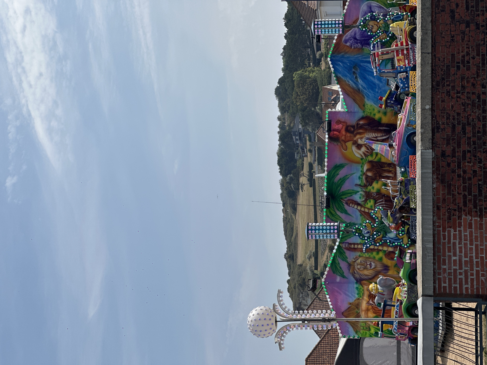 This image captures a colorful, amusement park ride set against a backdrop of a bright blue sky with a few scattered clouds. The ride features a vibrant mural depicting various animals and jungle scenes, including a lion, elephants, and a gorilla. Several small, rideable vehicles, designed to resemble cars, boats, and other whimsical transports, are positioned in front of the mural. These vehicles are painted in bright, contrasting colors and adorned with decorative lights.
To the left of the ride, a tall, spherical light fixture, covered in small bulbs, stands prominently. In the background, rolling green hills dotted with trees and houses suggest a semi-rural or suburban environment. The foreground shows a brick wall and a metal fence, indicating the observer's vantage point. The presence of the amusement ride suggests a public event or a permanent fairground. The sky is clear, with a few small birds visible in flight, and the overall lighting indicates it is daytime. The scene appears to be set in Hautot-sur-Mer, France, given the context of its location.