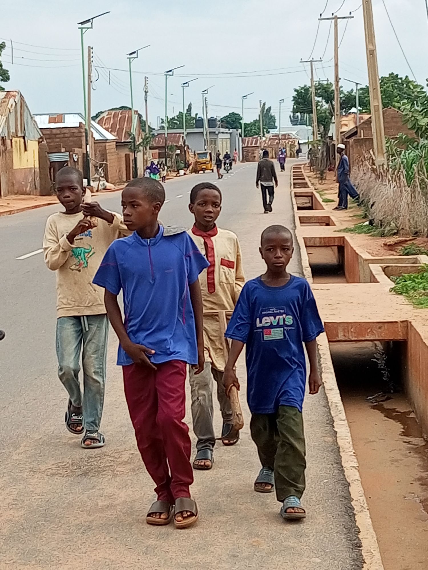 The image captures a street scene in Tempure, Nigeria, with five young boys walking on a paved road. The sky is overcast, suggesting a cloudy day. In the foreground, three boys are prominently featured, walking towards the viewer. The boy on the left wears a light-colored long-sleeved shirt with a graphic print and blue jeans, paired with dark sandals. The middle boy is dressed in a blue t-shirt with a zipper and maroon pants, also wearing dark sandals. The boy on the right is wearing a tan outfit with red trim and dark pants, with dark sandals. Behind them, two more boys are walking away from the viewer. The boy on the left, further back, is wearing a light-colored shirt and blue jeans. The boy on the right, slightly ahead, is dressed in a dark jacket and pants.
The environment features buildings with corrugated metal roofs on either side of the road, indicative of a local settlement. Utility poles with wires crisscross the scene, and some poles are equipped with solar-powered streetlights, a characteristic feature of areas with improved infrastructure. A drainage channel runs alongside the road on the right, with visible concrete structures. In the background, a yellow vehicle is parked, and other people and vehicles are present, suggesting a moderately active urban environment. The overall impression is a snapshot of daily life in a Nigerian town, with a blend of traditional housing and modern amenities.