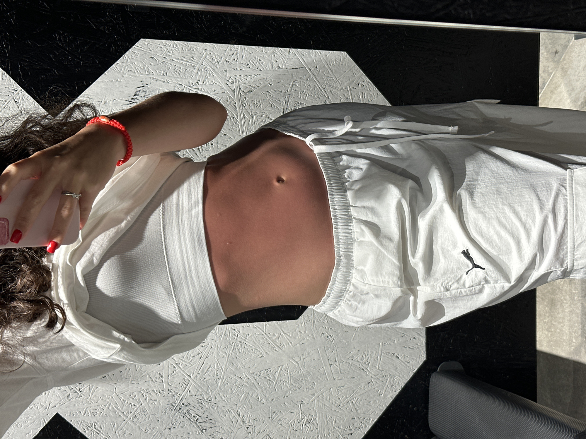 A woman lies on a black and white patterned surface, wearing a white crop top and white Puma sweatpants. She holds a red phone in her hand, her fingernails painted red. A red bracelet is on her wrist. The woman's abdomen is visible. The location is Pattaya City, Thailand.