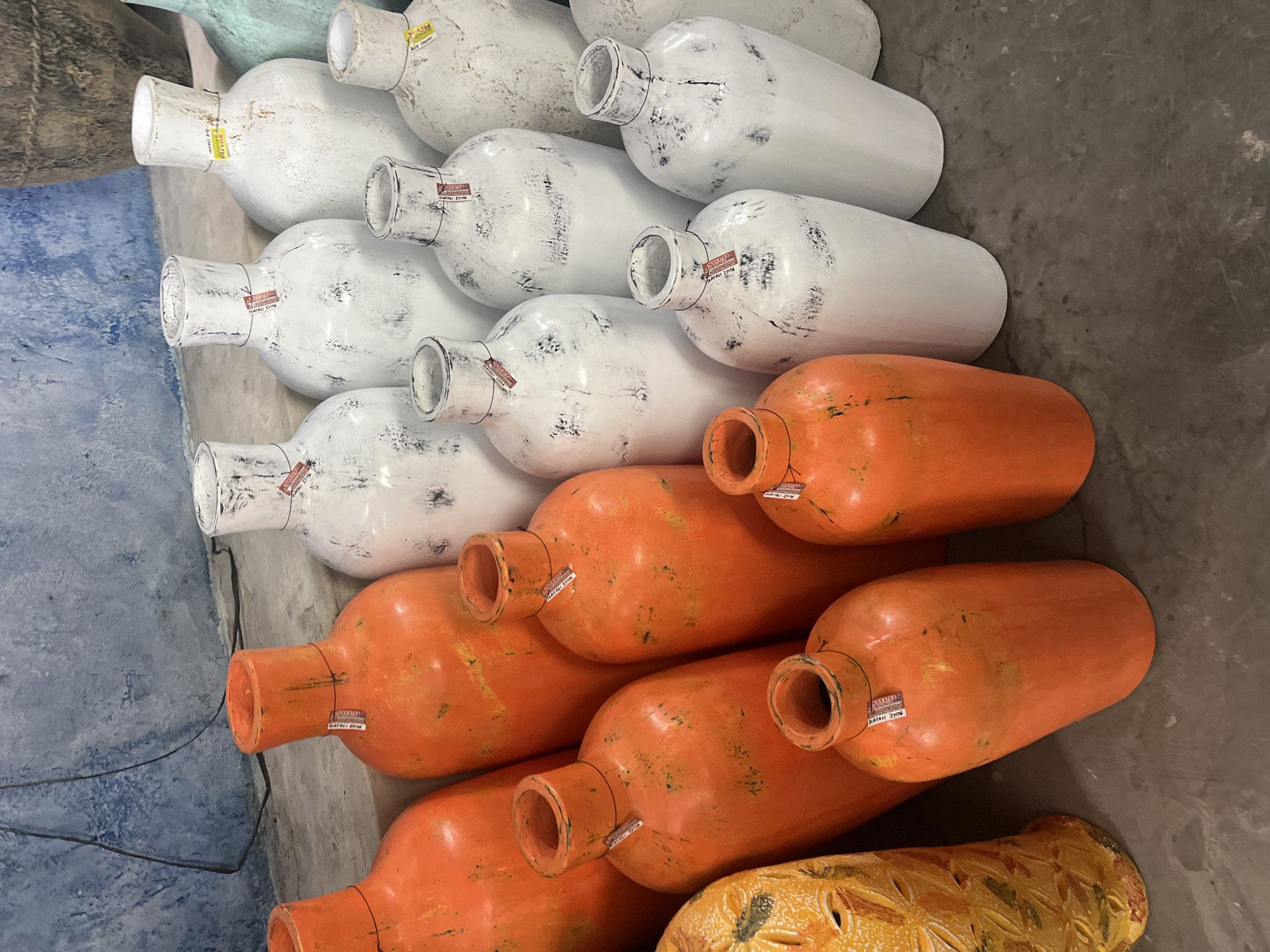A collection of ceramic vases is displayed on a concrete floor against a textured blue wall. The vases vary in color and size, with many in shades of white and orange, and a few in a muted green. Some of the white vases have a distressed, speckled finish, while the orange vases are a solid, bright hue. Several of the vases are decorated with small, red and white tags. To the left, a tall, yellow vase with intricate carvings is also visible. The overall impression is of a retail display of decorative pottery.