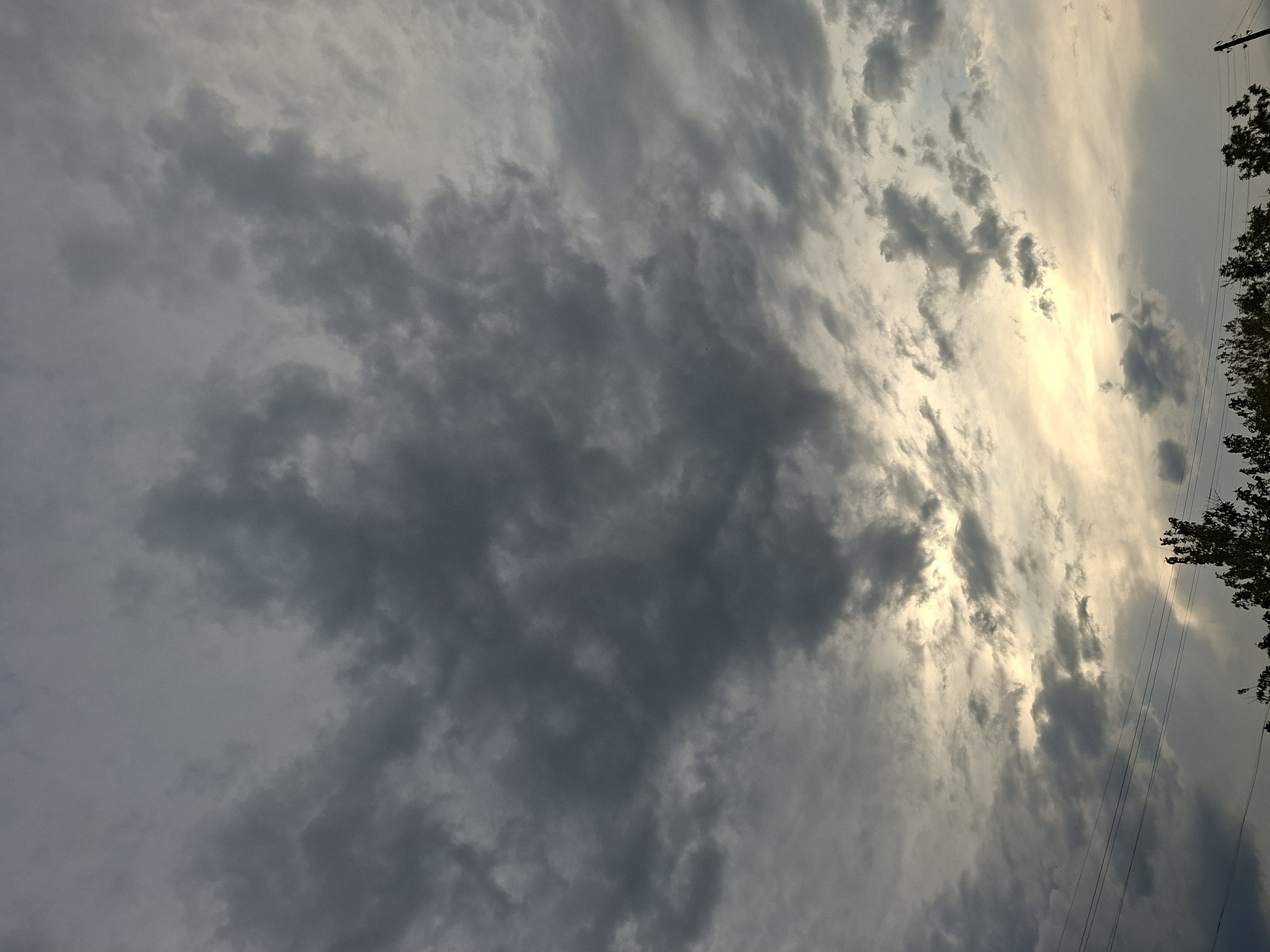 The image shows a cloudy sky. The clouds are predominantly gray and white, with some areas appearing darker, suggesting potential rain. A patch of brighter, yellowish light breaks through the clouds, hinting at the presence of the sun. Some tree branches are visible on the right side of the image, along with power lines. The image gives a low-angle view, looking upwards. The location of the image is given as Galabovo, Bulgaria.