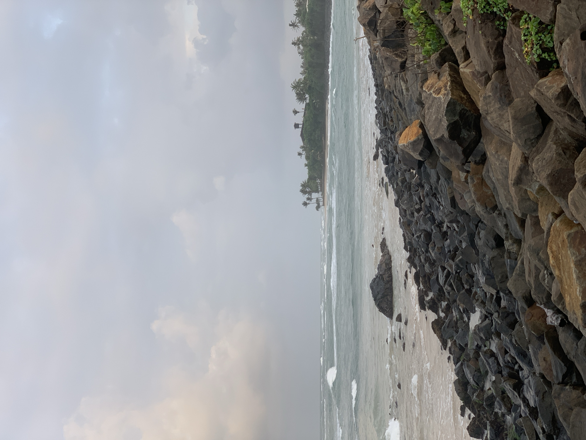 A stormy, overcast day at a rocky beach in Kathaluwa, Sri Lanka. The foreground is dominated by a dense collection of dark, jagged rocks, with a few larger, lighter-colored boulders interspersed. Beyond the rocks, a sandy shore meets the turbulent ocean. The sea is a muted teal color, with white-capped waves crashing against the shore and breaking further out. In the distance, a lush green peninsula juts out into the water, dotted with palm trees and a small, elevated structure resembling a watchtower. The sky is a uniform gray, with hints of pale yellow and pink near the horizon, suggesting either dawn or dusk. There are no people visible in the image.
