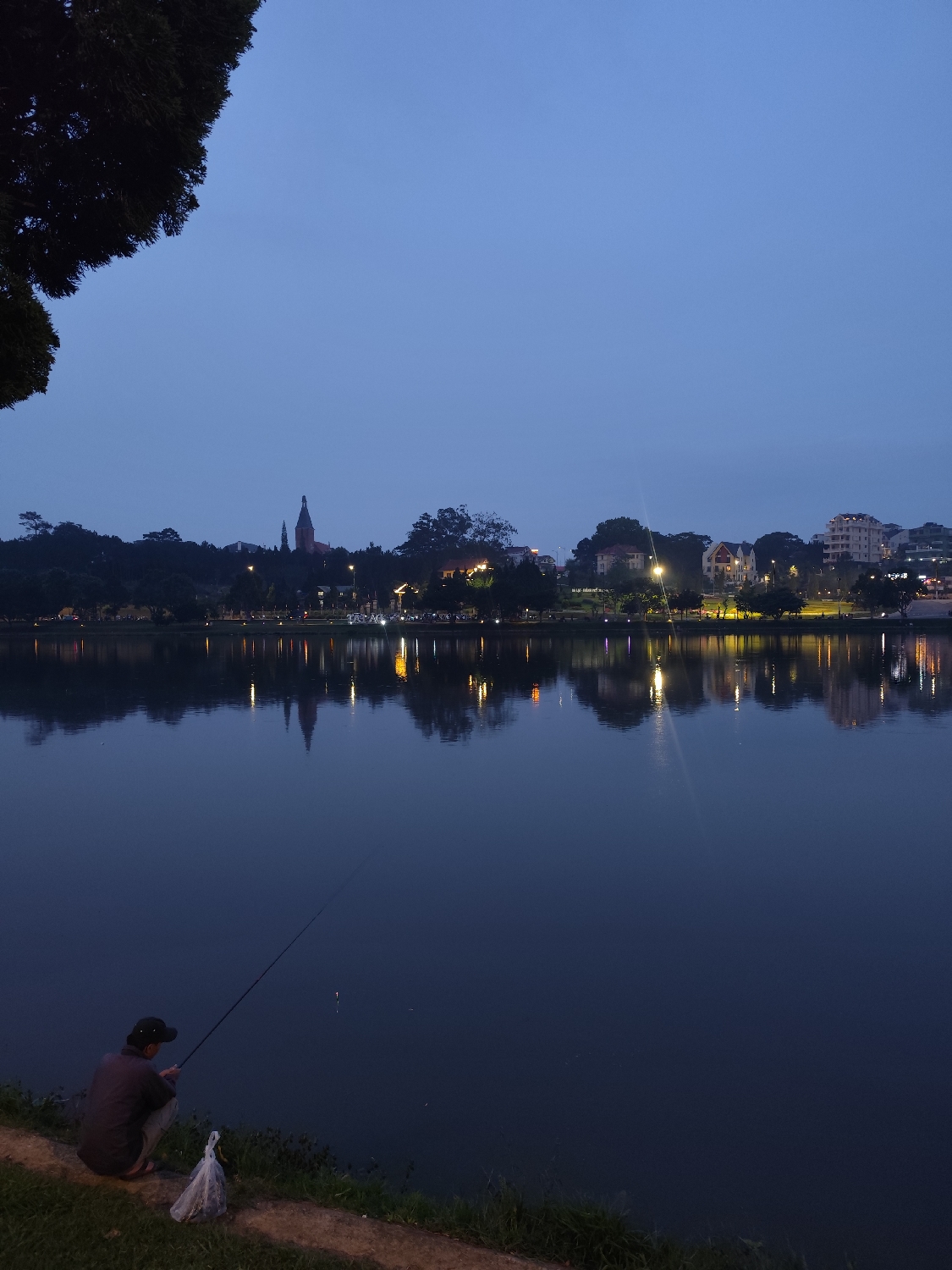 This media file captures a serene evening scene at Xuân Hương Lake in Đà Lạt, Vietnam. The primary subject is a lone individual, seen from behind, squatting on the grassy bank in the foreground. They are wearing a dark shirt and a dark baseball cap, actively engaged in fishing with a long rod extending into the tranquil lake. A white plastic bag rests beside them on the bank.
The lake, which dominates the midground, is exceptionally calm, perfectly reflecting the artificial lights and structures from the distant city shore. In the background, the silhouette of a city unfolds under a twilight sky. Various buildings, including residential structures and what appears to be a distinctive church steeple on the left, are nestled among dense trees. Numerous warm artificial lights illuminate the distant bank, casting vibrant, elongated reflections across the water's surface, creating a picturesque glow.
The sky is a deep, uniform blue, indicating dusk or early evening, with no visible clouds. A dark, leafy tree branch hangs into the frame from the upper left corner, partially obscuring the sky. The overall atmosphere conveyed is peaceful and quiet, characteristic of an evening spent fishing. No visible text is present in the image.