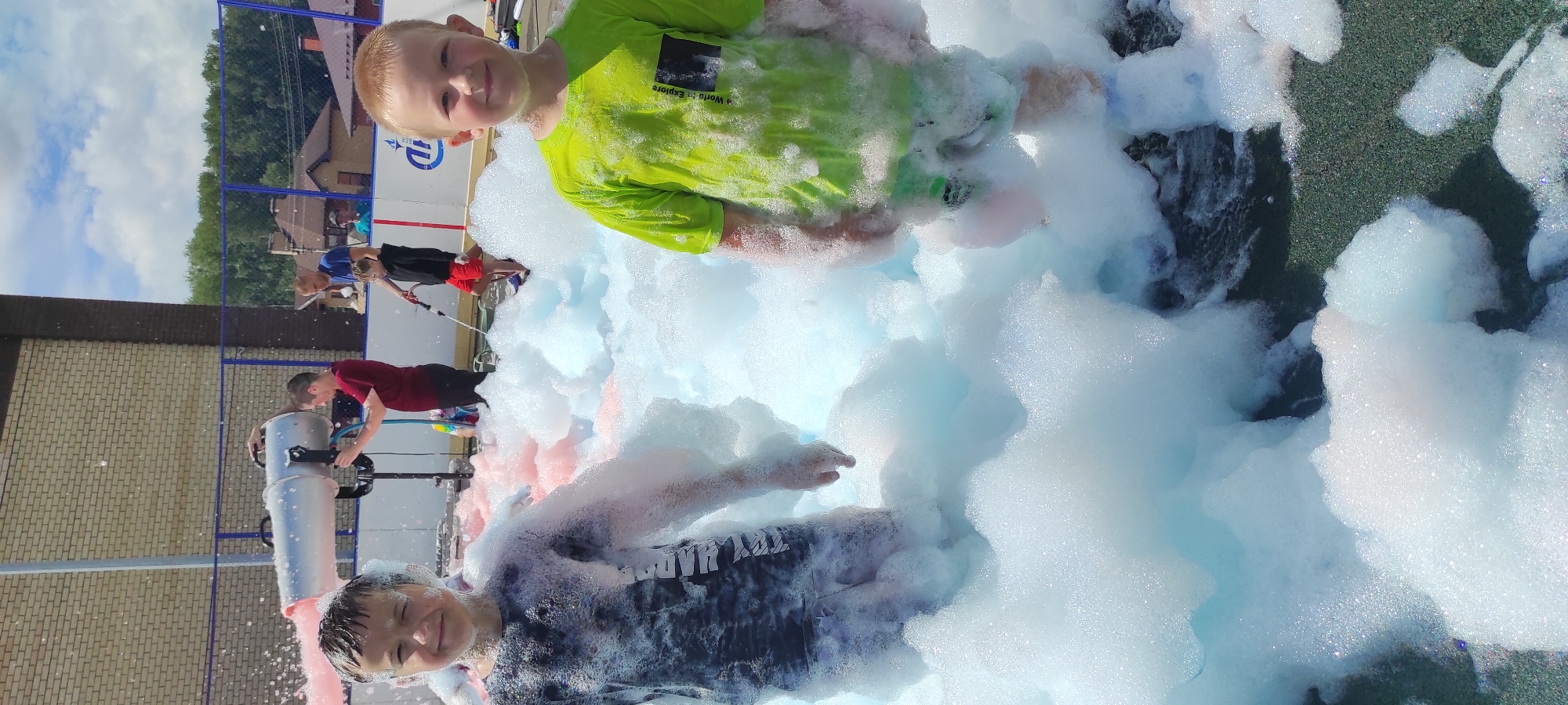 In Noviy, Russia, two young boys stand in a shallow pool of blue and pink colored foam, presumably at an outdoor event. The boy on the left, wearing a dark t-shirt with a white graphic, is partially obscured by the foam, which reaches his chest. He smiles and squints his left eye. The boy on the right, in a bright green t-shirt with a dark square graphic on the left chest, is also immersed in foam up to his knees. He looks directly at the camera with a slight smile. In the background, a foam machine is visible, operated by an adult wearing a maroon t-shirt. Another adult in a blue t-shirt and a child in a black hoodie and red shorts are also present, along with other onlookers further in the background. The environment appears to be an outdoor recreational area with a brick building and fencing visible behind the participants. The sky is partly cloudy.
