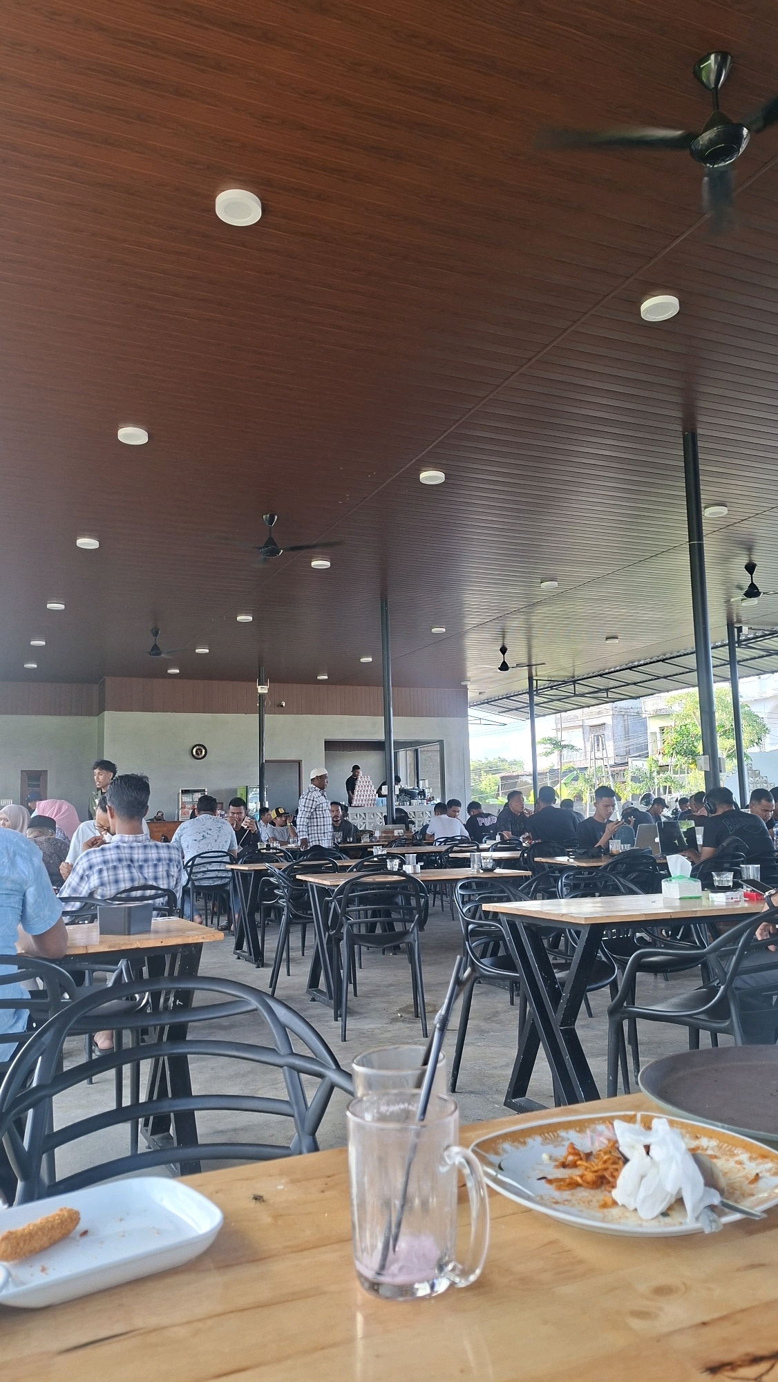 The image depicts an indoor-outdoor dining area of a restaurant, likely a cafe or eatery, located in Keudebayu, Indonesia. The ceiling is constructed of dark wood panels and features recessed lighting and several ceiling fans, suggesting a warm climate. The dining area is populated with numerous tables and black chairs, indicating it is a busy establishment. In the foreground, a wooden table is visible, set with a white rectangular plate containing a single fried food item, a clear glass mug with a pink beverage and two black straws, and a partially visible plate with used napkins and remnants of food.
The background reveals a semi-open-air setting, with a light-colored roof structure supported by dark poles. Beyond this, trees and buildings are visible under a bright sky, suggesting the location is an urban or semi-urban area.
Several individuals are present throughout the scene, engaged in dining and conversation. In the mid-ground, groups of people are seated at tables, some appearing to be eating or looking at their phones. A few individuals are standing and interacting with others. The overall atmosphere appears to be casual and social. The architectural style, with the combination of wood and open spaces, is consistent with modern Indonesian design for such establishments.