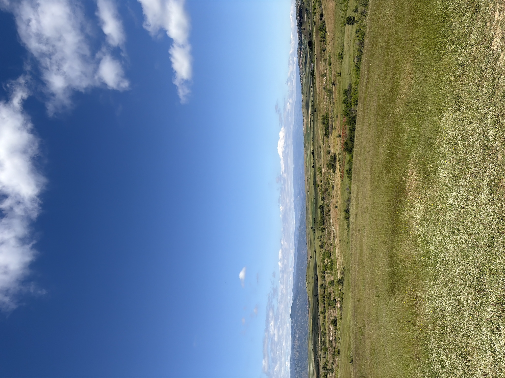 The image depicts a landscape with a clear blue sky, scattered white clouds, and a view of grassy terrain with sparse trees and shrubs. In the distance, a mountain range is visible. The photograph appears to be taken in an outdoor setting, showing natural elements such as sky, clouds, mountains, grass, and trees. No people or animals are immediately visible. The location and city are Unknown.