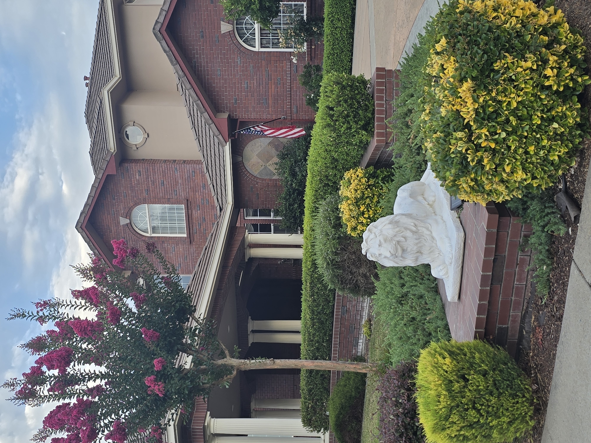 A white lion statue sits on a brick platform in front of a large, red brick house with a multi-level roof. A Crape Myrtle tree with pink flowers is to the left of the house. The house has a covered porch supported by white columns and several arched windows. An American flag hangs from a pole near the front of the house. The yard is landscaped with manicured green hedges and decorative bushes. The sky is cloudy.