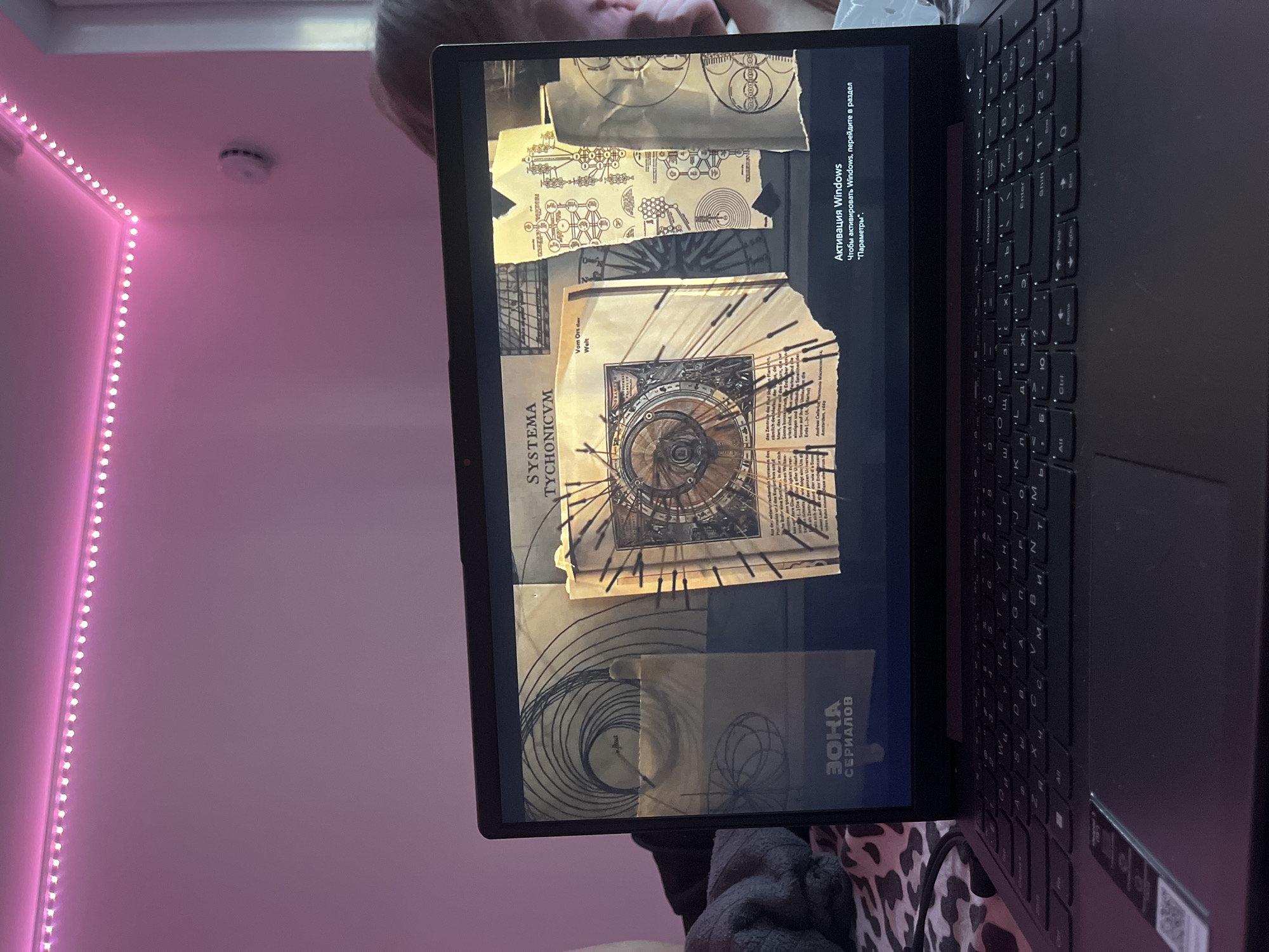 The image depicts a laptop displaying a collection of images, including diagrams labeled "SYSTEMA TYCHONICVM" and "ZOHA CEPUANOB" in Russian, along with a Windows activation message in Russian. A keyboard is visible at the right edge of the laptop. The background includes a wall with pink LED strip lights and a smoke detector. The location appears to be indoors. The city is not visible, so a location within Saint Petersburg, Russia cannot be confirmed.