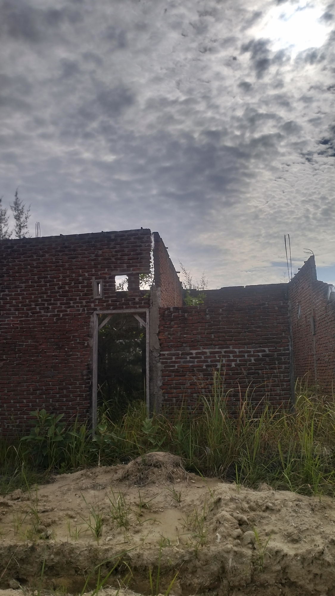 The image depicts an unfinished, partially constructed building made of red brick, located in Lhokseumawe, Indonesia. The structure appears abandoned, with exposed brickwork and incomplete walls. A doorway is visible, leading into a dark, overgrown interior filled with vegetation. The foreground is dominated by a mound of disturbed earth and scattered grass. The sky above is overcast with textured clouds, with a bright spot indicating the sun's presence behind the clouds. Several thin, reedy plants are visible on the left side of the frame, and taller grasses grow around the base of the building. The overall scene suggests a construction site that has been left undeveloped, blending with the natural environment.