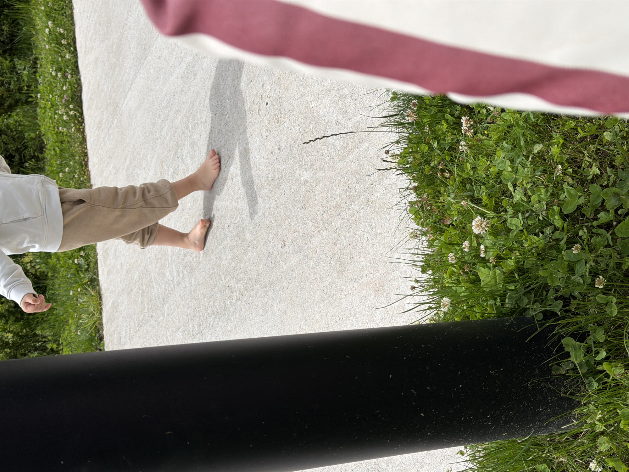 A child in tan pants and a white top stands barefoot on a concrete surface next to grass. The lower portion of a black cylindrical object is visible. A red and white striped fabric is in the background. The location is speculated to be Chkalov, Armenia.