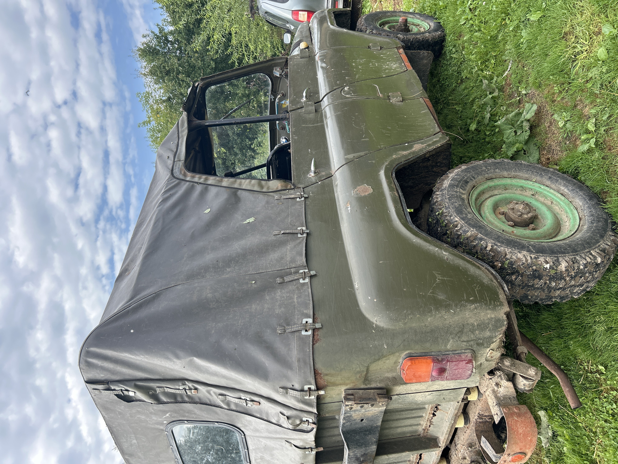 In the image, a weathered, olive-drab UAZ-469 off-road vehicle is parked on a grassy area, likely in or around the village of Kalitsino, Russia. The vehicle's canvas top is darkened and appears worn, with visible straps and buckles securing it. Rust spots are present on the body of the vehicle, particularly on the rear fender and near the top canvas attachment points. The rear taillight assembly is visible, and the vehicle has large, muddy tires with green rims. A portion of another vehicle, possibly a silver SUV, is partially visible to the right of the UAZ. The environment suggests an outdoor setting with green grass and a backdrop of trees and a cloudy sky. There are no people or animals present in the scene, and no specific actions are being depicted beyond the vehicle being stationary. The overall impression is of a utilitarian vehicle, possibly aged or used for off-road purposes, in a rural or semi-rural location.