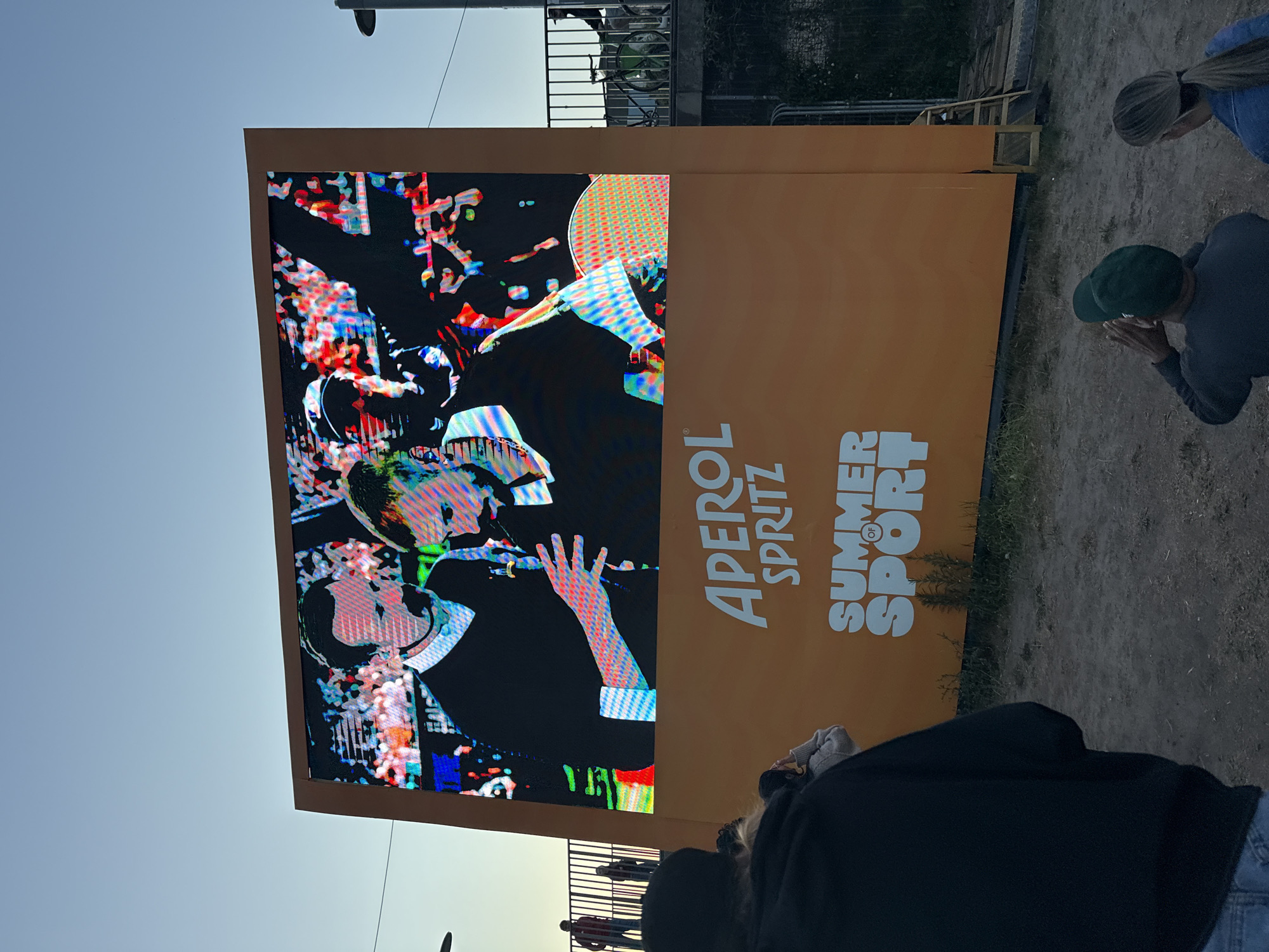 A large outdoor LED screen displays a colorful image of two men on stage, one holding a microphone. Below the image, the words "APEROL SPRITZ SUMMER OF SPORT" are printed in large white letters on an orange background. Several people are visible in the foreground, some watching the screen, others looking away. The ground is dirt and gravel. The sky is clear and blue.