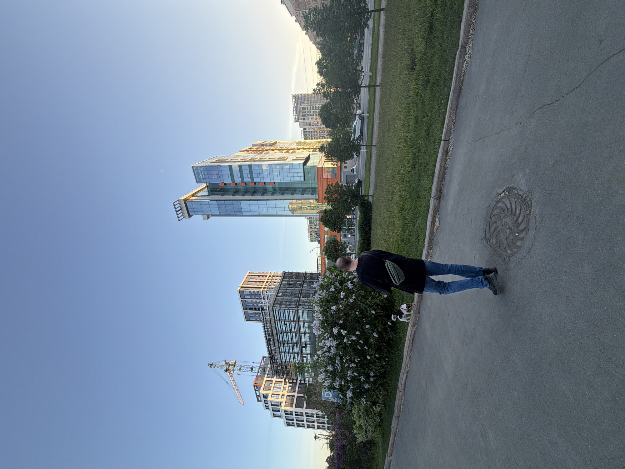 The image depicts a person standing on a sidewalk next to a park in a city, potentially Izhevsk, Russia. The person is wearing jeans, a dark jacket, and dark shoes, and they appear to be holding the leash of a small dog. Tall modern buildings are visible in the background, with a construction crane next to one. Lush greenery is along the walkway and includes flowering bushes. A manhole cover is visible on the sidewalk. The sky is blue.