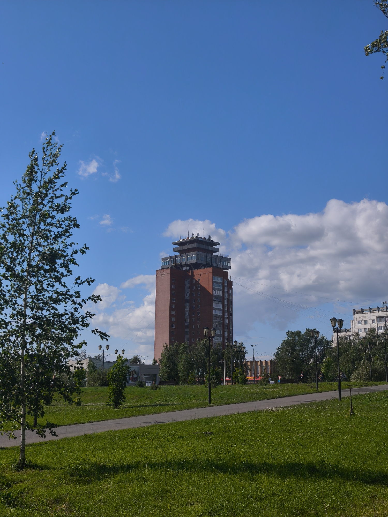 This is a photograph of the city of Yoshkar-Ola, Russia, depicting a park with a distinctive brick high-rise building in the background. The building is multi-story with a unique, multi-tiered structure at its summit. A paved pathway winds through a lush green park in the foreground, with well-maintained grass and scattered trees. The sky is a bright blue with scattered white clouds. Several lampposts are visible along the pathway, and in the distance, other buildings and trees can be seen. The scene suggests a pleasant day in an urban park.