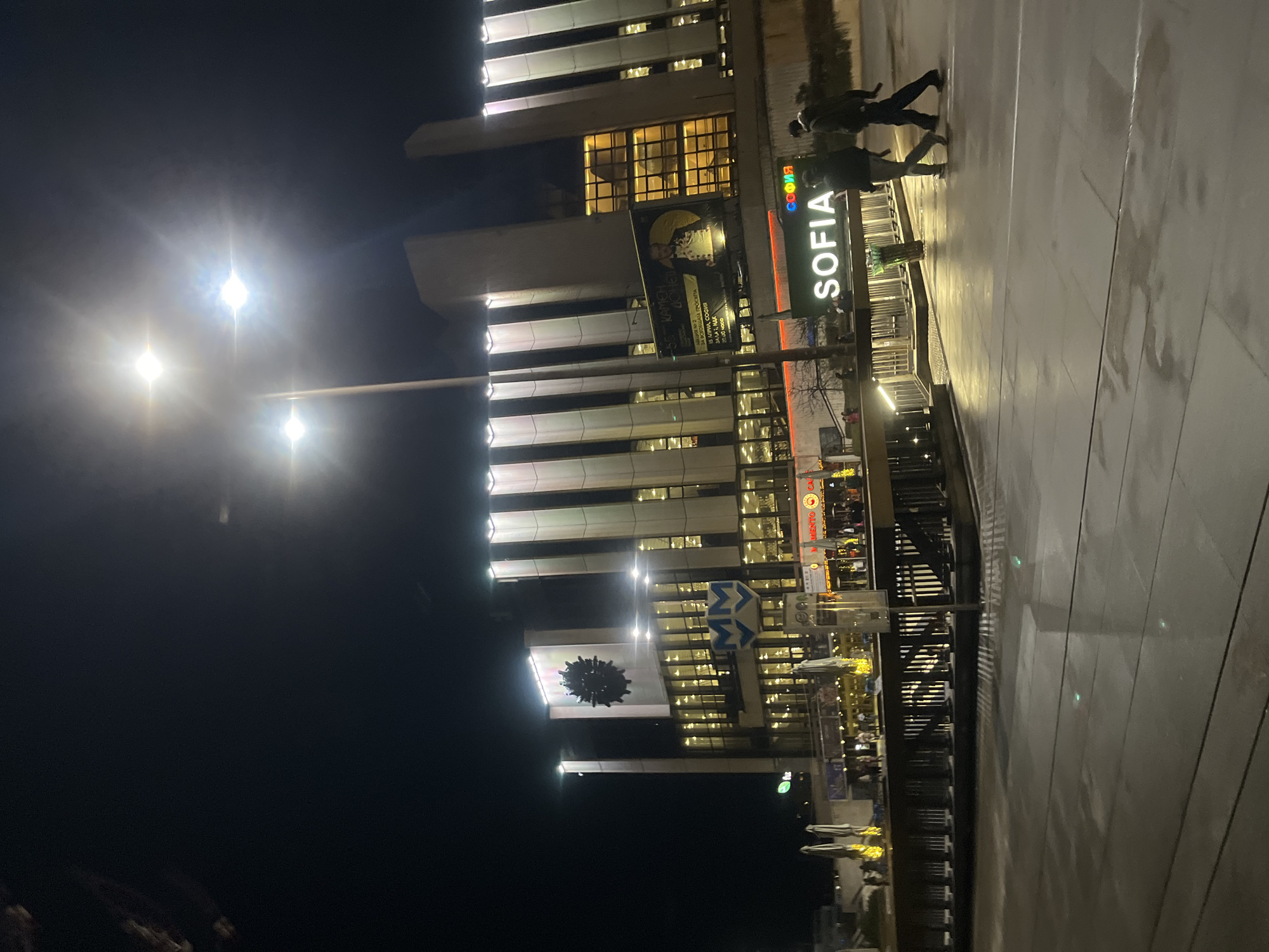 The image shows a nighttime view of Sofia, Bulgaria. The architecture of the city is visible through tall buildings with modern designs illuminated with lights. A sign reads "SOFIA" in large letters, indicating the location. A banner promoting "KAMEH" event can be seen on one of the buildings. Pedestrians are walking on a wet pavement. A sign of "MM" is visible next to an ornament shaped like a sun.