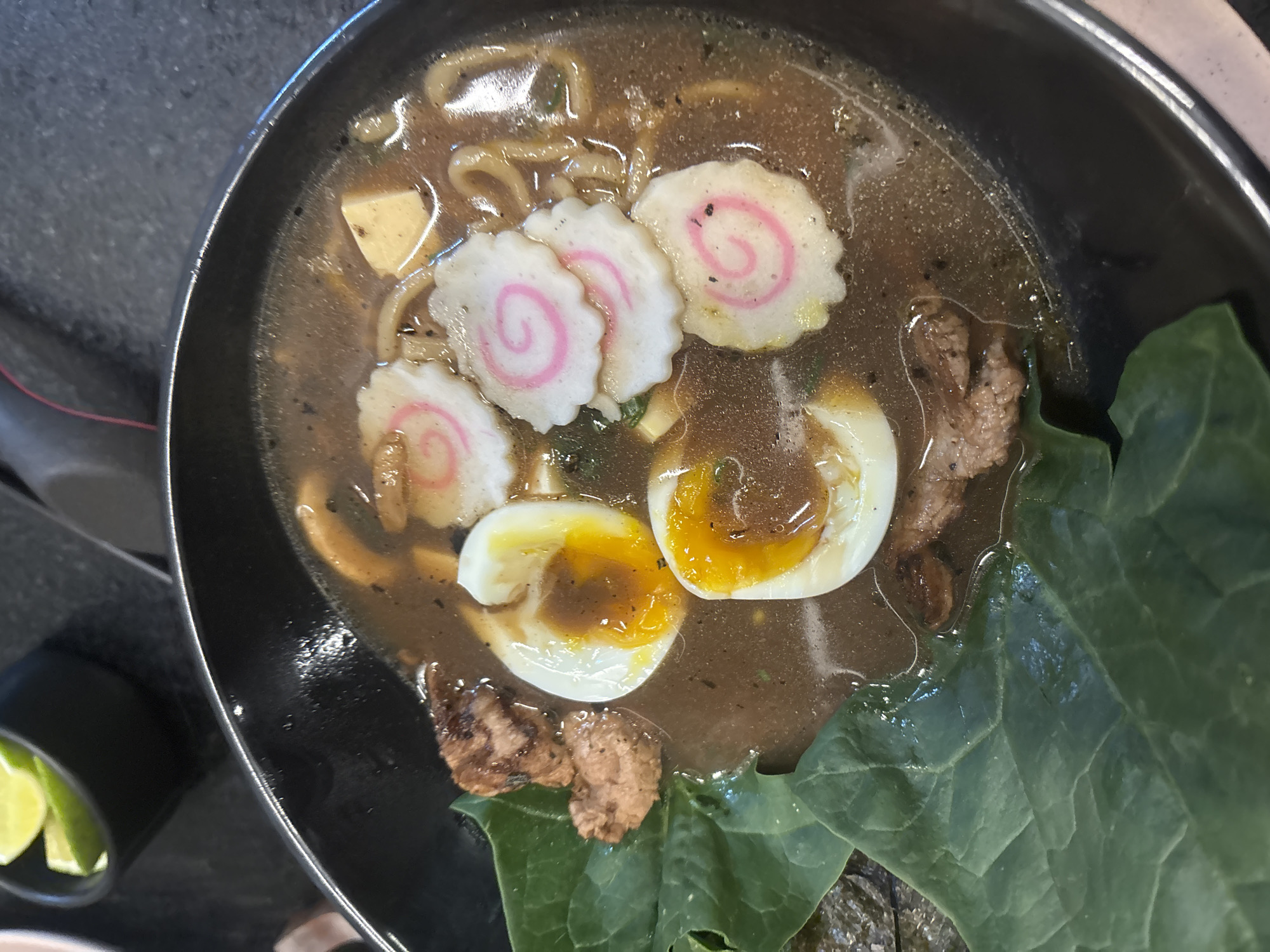 A close-up, high-angle shot presents a bowl of ramen, likely located somewhere other than the city of San Bernardino Tlaxcalancingo, Mexico. The ramen consists of noodles submerged in a dark broth, topped with sliced kamaboko (fish cake) featuring a pink swirl, halved soft-boiled eggs with runny yolks, pieces of meat, tofu, and leafy greens. A small dish containing lime wedges is visible in the bottom left corner of the image. The bowl itself appears to be black. The surrounding surface is out of focus.