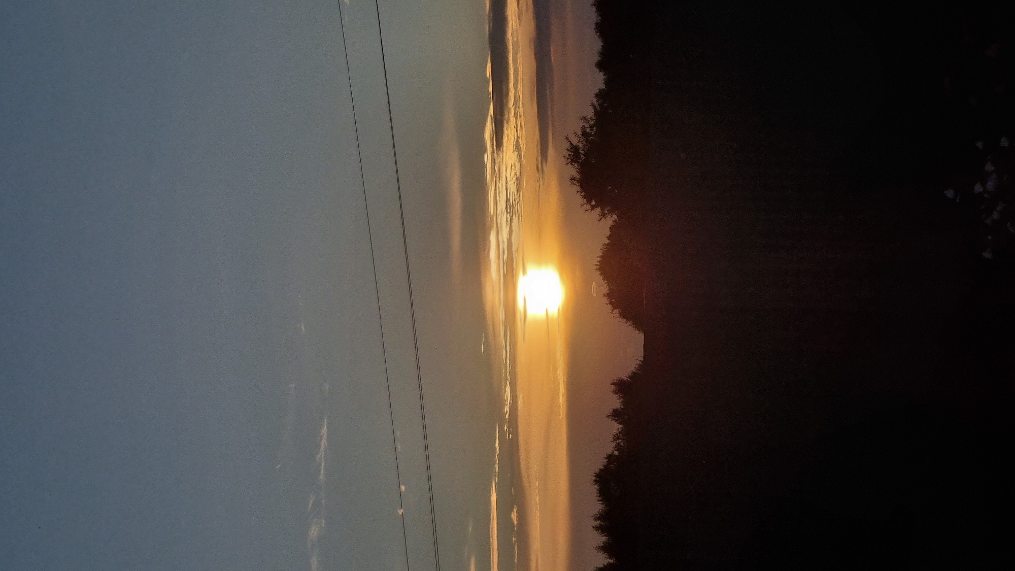 The image captures a sunset over a silhouette of trees and a field, likely located in or near Vysokoe, Russia. The sun is a bright, glowing orb partially obscured by a horizontal band of clouds, casting a warm orange and yellow light across the sky. Above the sun, the sky transitions from a pale orange to a dusky blue, with thin, wispy clouds scattered throughout. Two dark, horizontal lines, possibly power lines, stretch across the upper portion of the frame. The foreground is dominated by the dark silhouettes of trees and what appears to be a field, with the very bottom edge of the image showing indistinct, scattered light-colored objects, possibly debris or reflective surfaces on the ground. The overall impression is one of a tranquil evening scene.