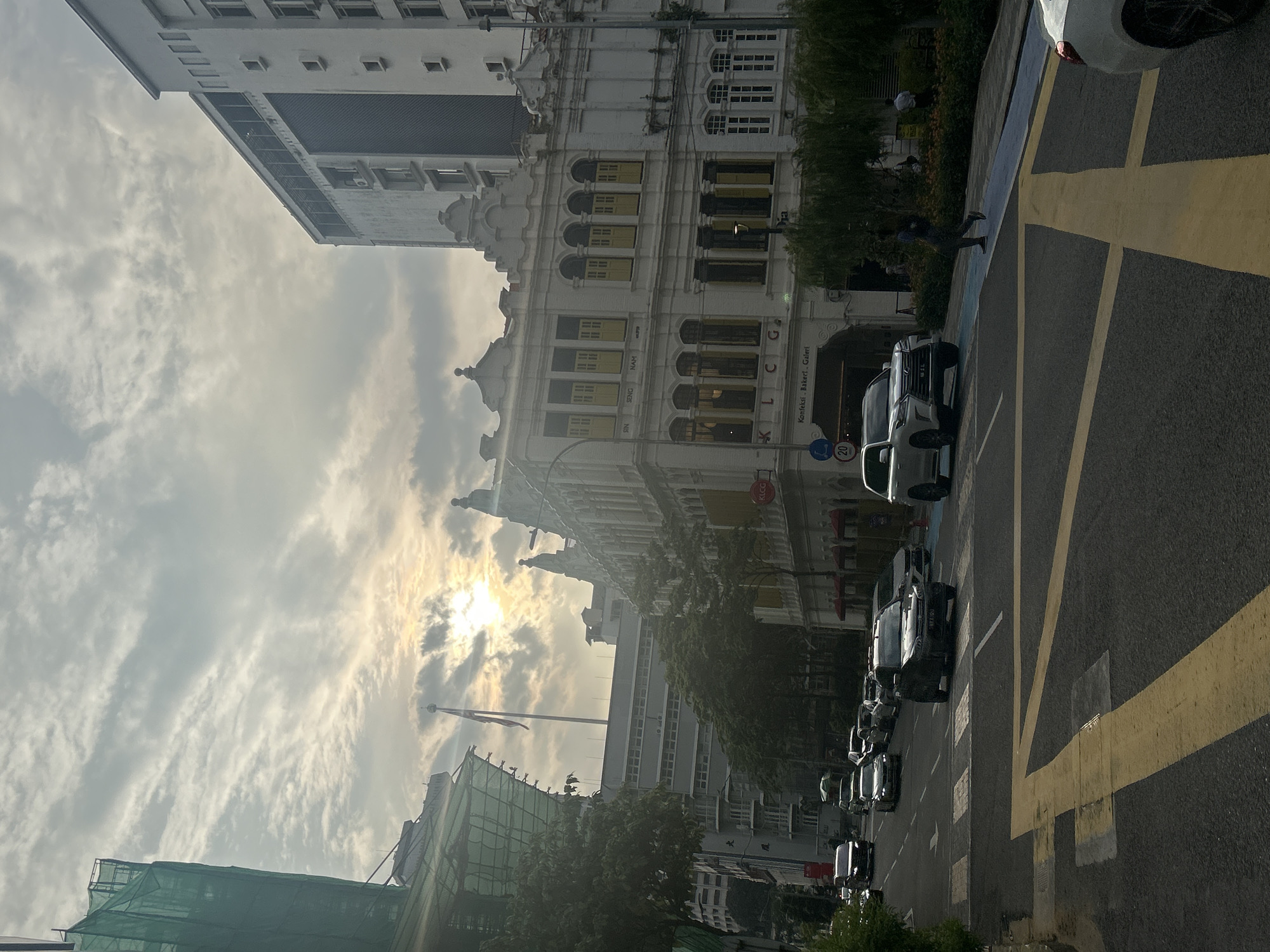 This image captures a street scene in Kuala Lumpur, Malaysia, likely in the vicinity of Jalan Sultan Ismail, given the architectural style and the presence of KLCC signage visible on a building. The time of day appears to be late afternoon or early morning, with the sun casting a warm glow through cloudy skies and illuminating the scene from the upper left quadrant.
In the foreground, a road with yellow and white road markings, including a pedestrian crossing, occupies the lower portion of the frame. Several vehicles are present on the road. A black SUV is stopped at the pedestrian crossing, with a white SUV behind it. Further down the street, a line of cars is moving.
Dominating the middle ground is a row of traditional colonial-era buildings with ornate white facades and yellow shutters. One building has a sign that partially reads "KLCC Konfeksi Bakery Galeri". Trees with lush green foliage line the sidewalk in front of these buildings.
In the background, a modern, tall white building stands to the right. To the left, a building under construction is visible, draped in green netting. A flagpole with a flag is also prominent in the mid-ground, against the sky.
A few individuals are discernible in the scene. One person, dressed in dark clothing, is walking on the sidewalk near the building with the "KLCC" sign. Another person, in a light-colored shirt and dark pants, stands further down the sidewalk.
The overall atmosphere is one of urban activity with a blend of historical and modern architecture, under a dramatic, cloud-filled sky.