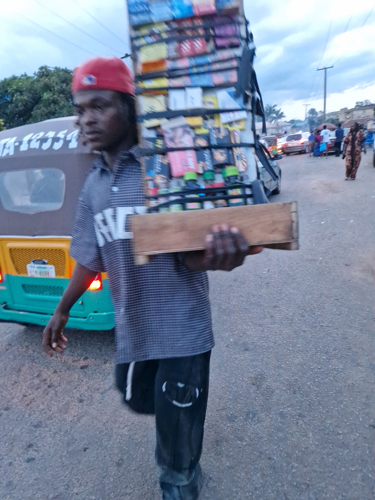 A young African male, wearing a red cap and a checkered shirt, walks with a large stack of colorful goods balanced on his head. He is holding a wooden plank in his right hand, presumably to help with the load. To his left, a yellow and green auto-rickshaw, also known as a Keke Napep, is visible. The man is on a paved road, which appears to be a street in Jos, Nigeria, based on the presence of other vehicles and pedestrians in the background, characteristic of a bustling urban environment. The sky is overcast with clouds.