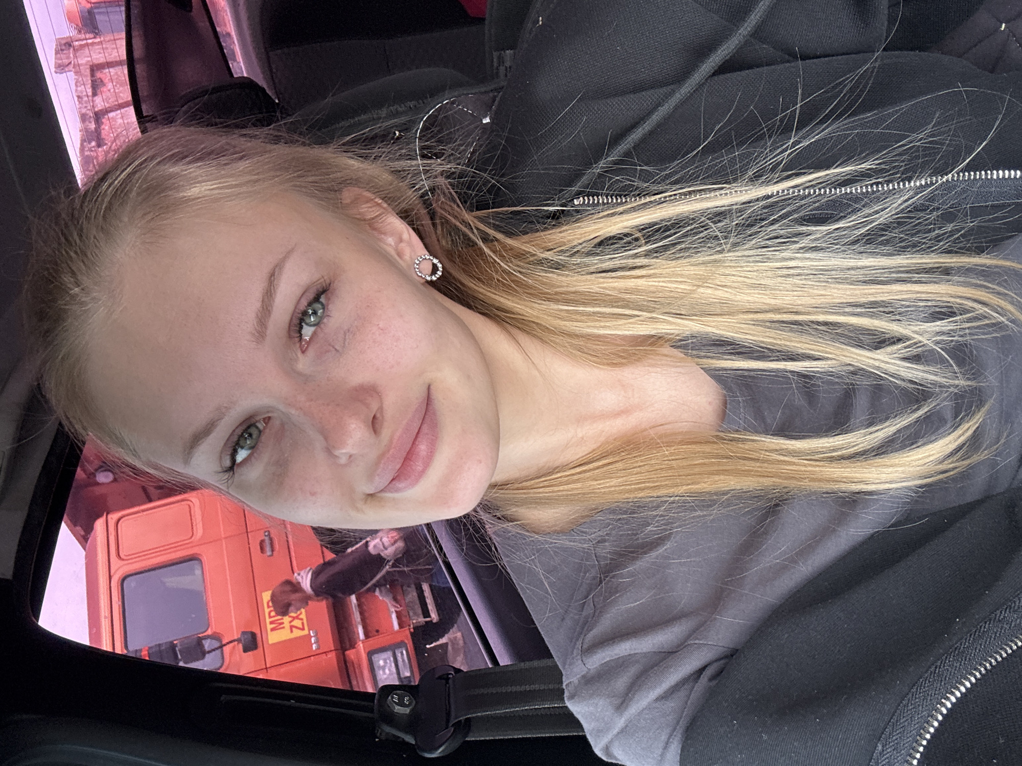 The image shows a young woman with blonde hair and blue eyes, wearing a gray t-shirt and a black zip-up hoodie. She is sitting in the driver's seat of a vehicle. Through the open car window, a large orange truck is visible, as well as another person walking in the background. The sky appears to be tinted pink, possibly due to the time of day or atmospheric conditions. The setting appears to be outdoors, possibly in a parking lot or along a road. There are no discernible landmarks that would specifically place this image in Ust'-Syny, Russia.