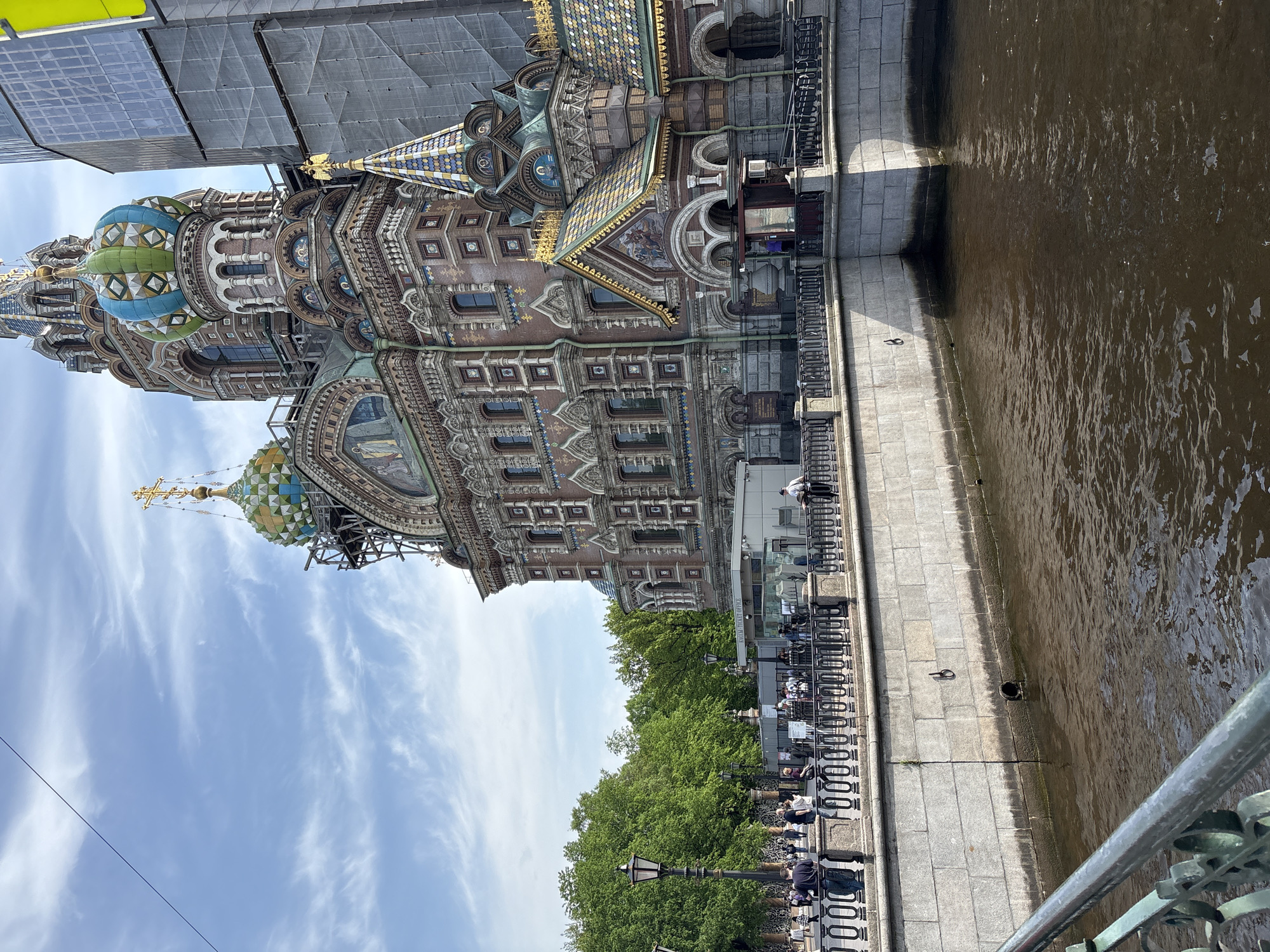The image shows the Church of the Savior on Spilled Blood in Saint Petersburg, Russia. The church is located on the embankment of the Griboyedov Canal. The building has multiple domes with colorful and intricate decorations. Scaffolding is visible around some of the domes, suggesting ongoing restoration work.
Alongside the canal, there's a stone embankment with a wrought-iron fence. People are sitting on benches and walking along the embankment. Trees are visible behind the embankment, adding greenery to the scene. The water in the canal is murky. The sky is partially cloudy with a light blue hue.