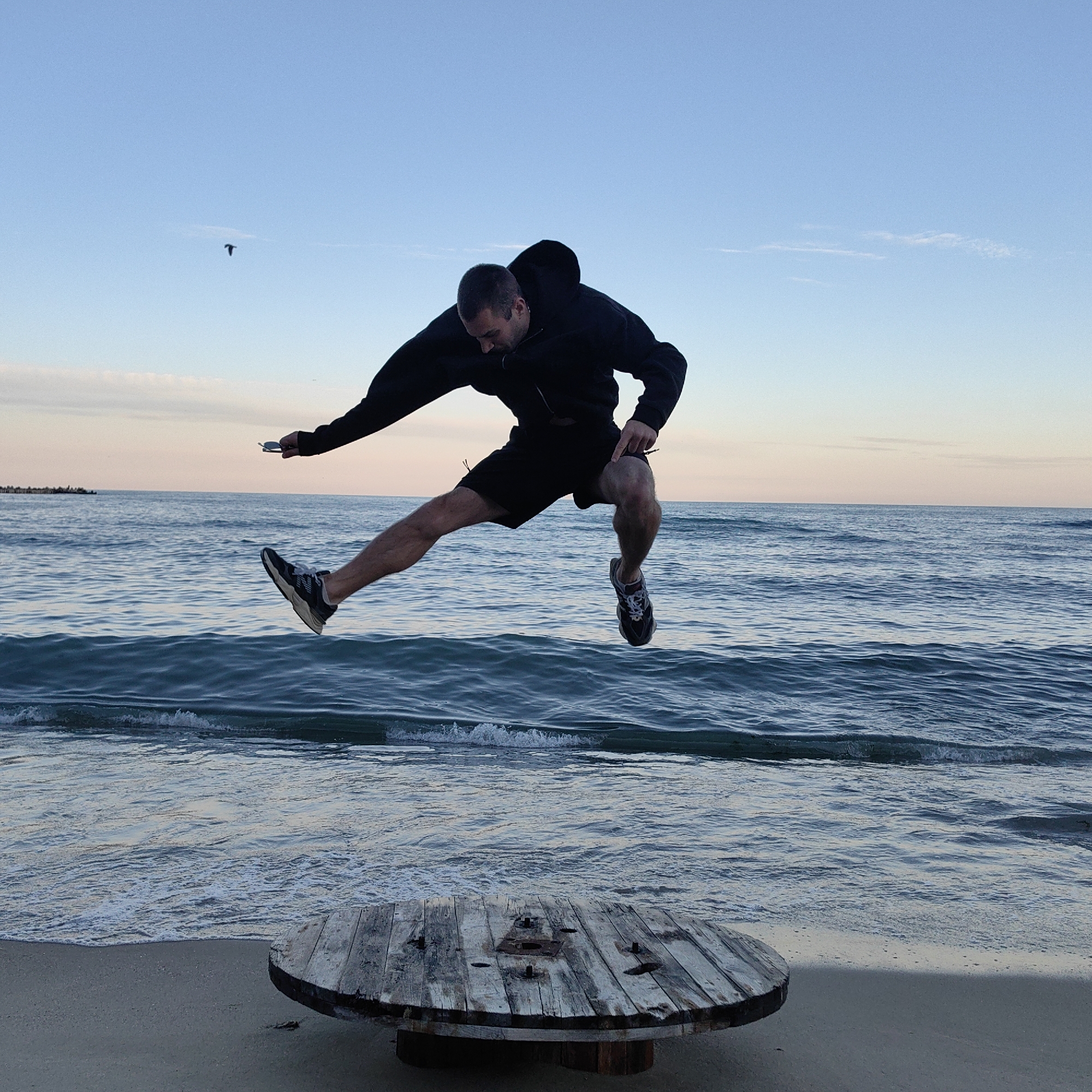 A man wearing a black hooded jacket and black shorts is leaping over a large, round wooden spool on a sandy beach. He's wearing athletic shoes and appears to be holding a small object in his left hand. In the background is the sea, with gentle waves and a faint pink and blue sky. A bird is visible in the sky. This photo was taken on the beach in Varna, Bulgaria.