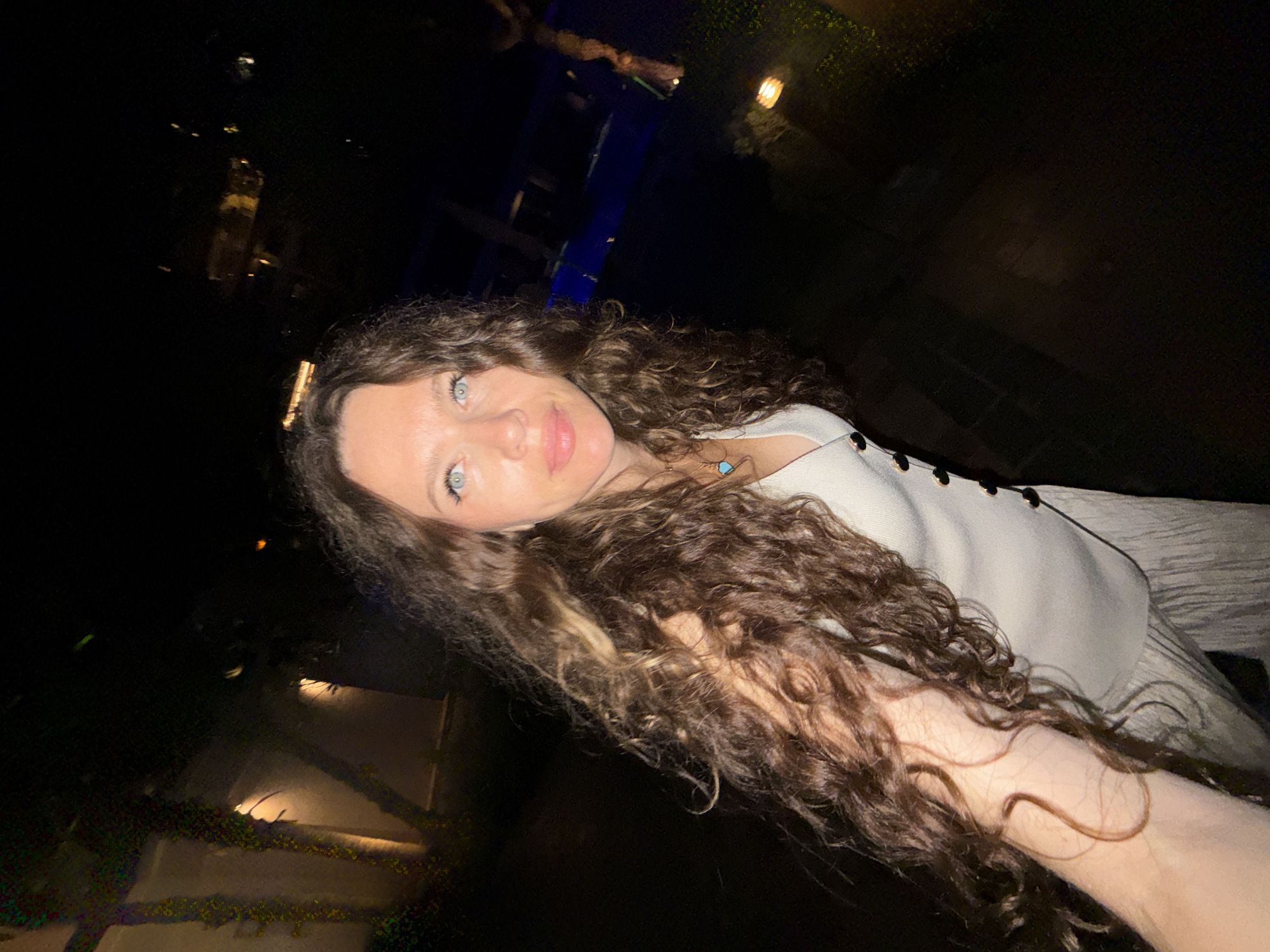 A woman with long, curly brown hair and blue eyes is posing for a selfie at night. She is wearing a light-colored sleeveless top and a light-colored skirt. She has a necklace with a turquoise pendant. The background appears to be an outdoor setting with some lighting, possibly a resort or a courtyard. The overall impression is that of a casual, nighttime portrait. There is no specific information about the location being in Dubai, United Arab Emirates, in the provided image.