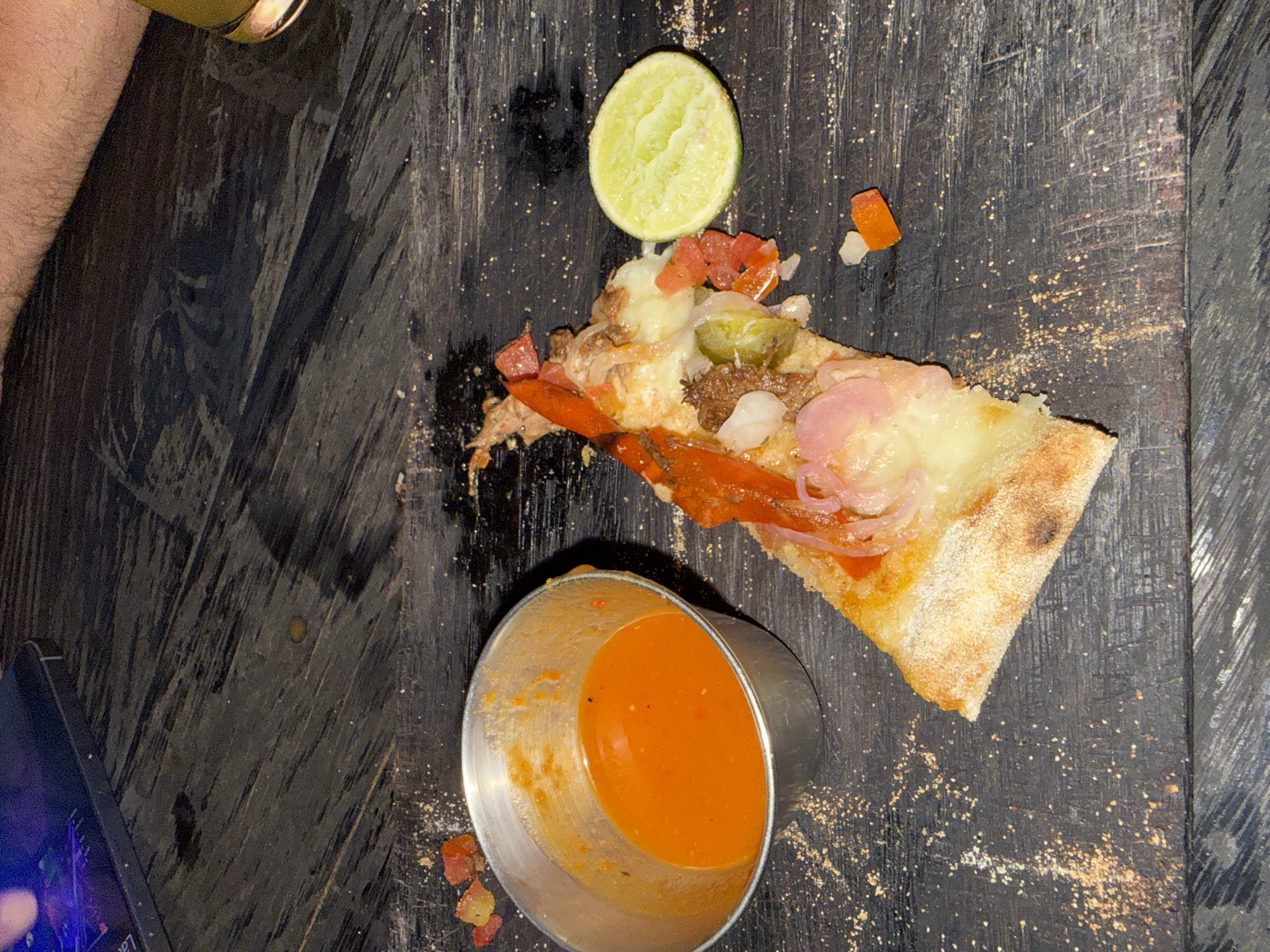 A partially consumed pizza slice rests on a dark, possibly wooden, table surface. The pizza is topped with cheese, red onions, tomatoes, peppers, and what appears to be meat. A lime wedge sits near the pizza. A small metal container holds an orange-colored sauce. Scattered around the pizza and sauce are crumbs and small pieces of food. The edge of a phone or tablet is visible in the lower left corner, and a person's arm is visible in the upper left. Based on prior knowledge, this image may be taken at a venue in North Kuta, Indonesia, known for pizza and sauce.