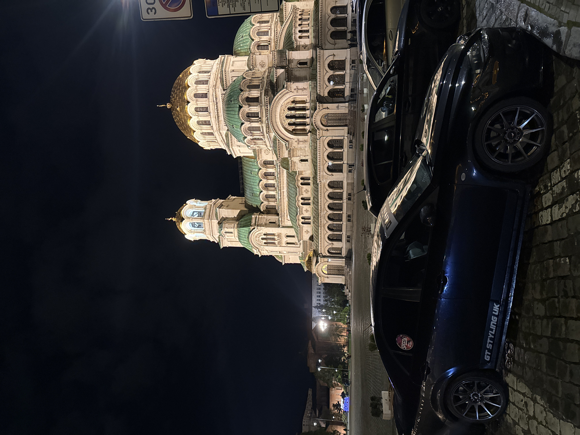 The image, taken at night, shows the illuminated Alexander Nevsky Cathedral in Sofia, Bulgaria. To the right, two dark-colored cars are parked on a cobblestone street. One car has "GT STYLING UK" written on its side. Streetlights illuminate the area beyond the cathedral. A street sign is visible in the upper left. The sky is dark.