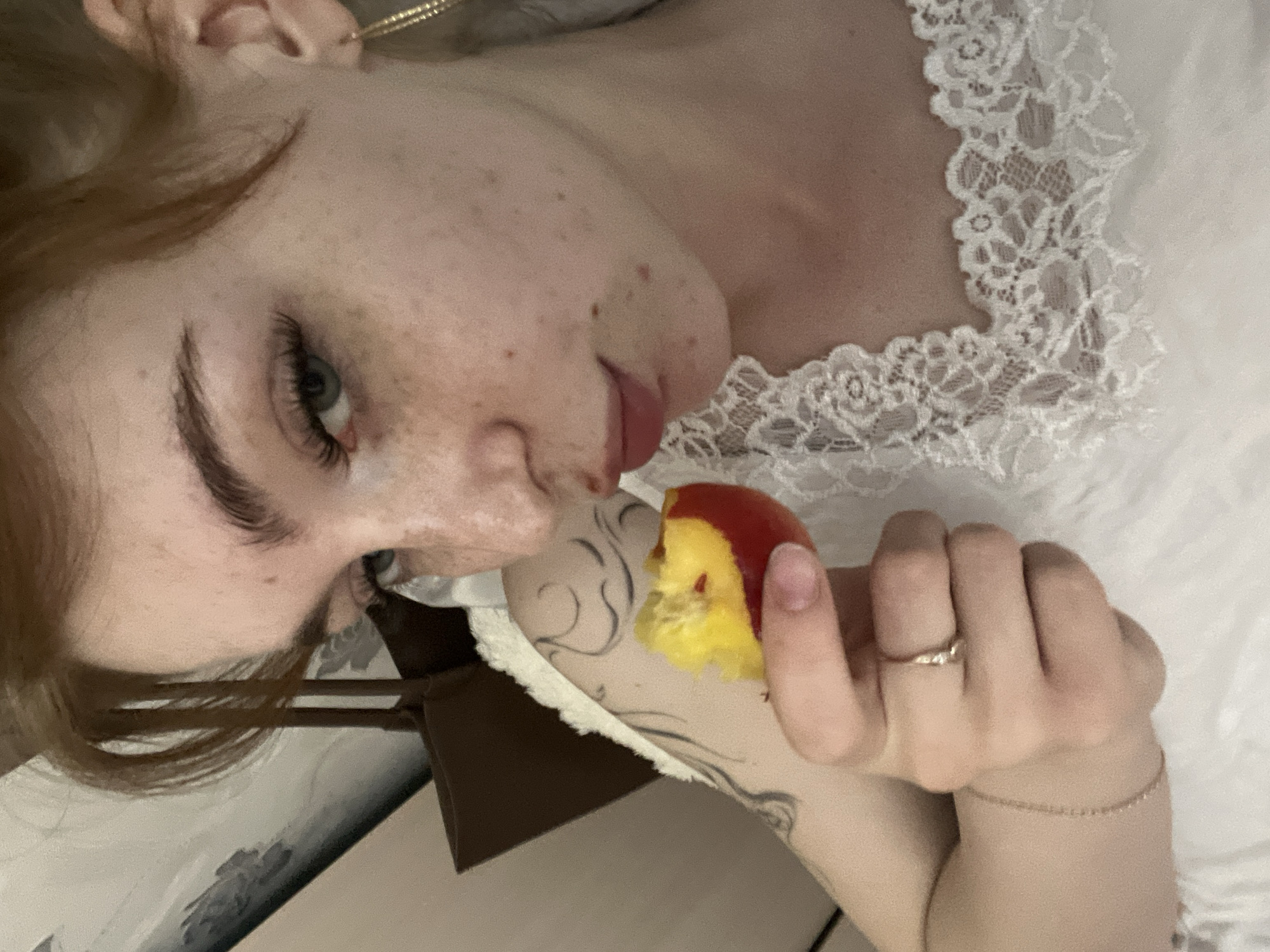 The image is a close-up, selfie-style shot of a young person with auburn hair and fair skin, lightly freckled. They are looking towards the camera with one eye visible, the other partially obscured by hair. The person is wearing makeup, including false eyelashes, and has a delicate gold hoop earring in their visible ear. They are holding a bite-sized piece of a peach or nectarine up to their mouth, appearing to be in the process of eating it. The person is wearing a white garment with lace trim. A tattoo is visible on their left arm, and a thin bracelet is on their left wrist. A ring is on their right index finger. The background appears to be an indoor setting, possibly a bedroom, with a textured wall and a hint of a brown bag or purse. The overall lighting is soft and diffused. There is no indication of the image being located in the city of Vetluzhsky, Russia.