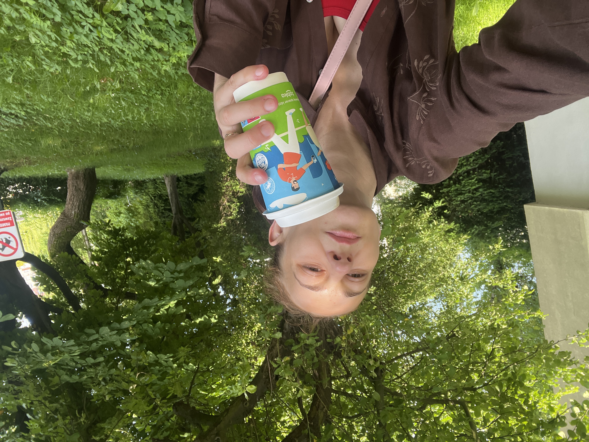 A woman in a brown shirt holds a coffee cup with a picture of a man and woman on it. She is sitting outside under a tree with green leaves. There is a sign in the background that is partially visible and has red lettering. The ground is covered in green grass.