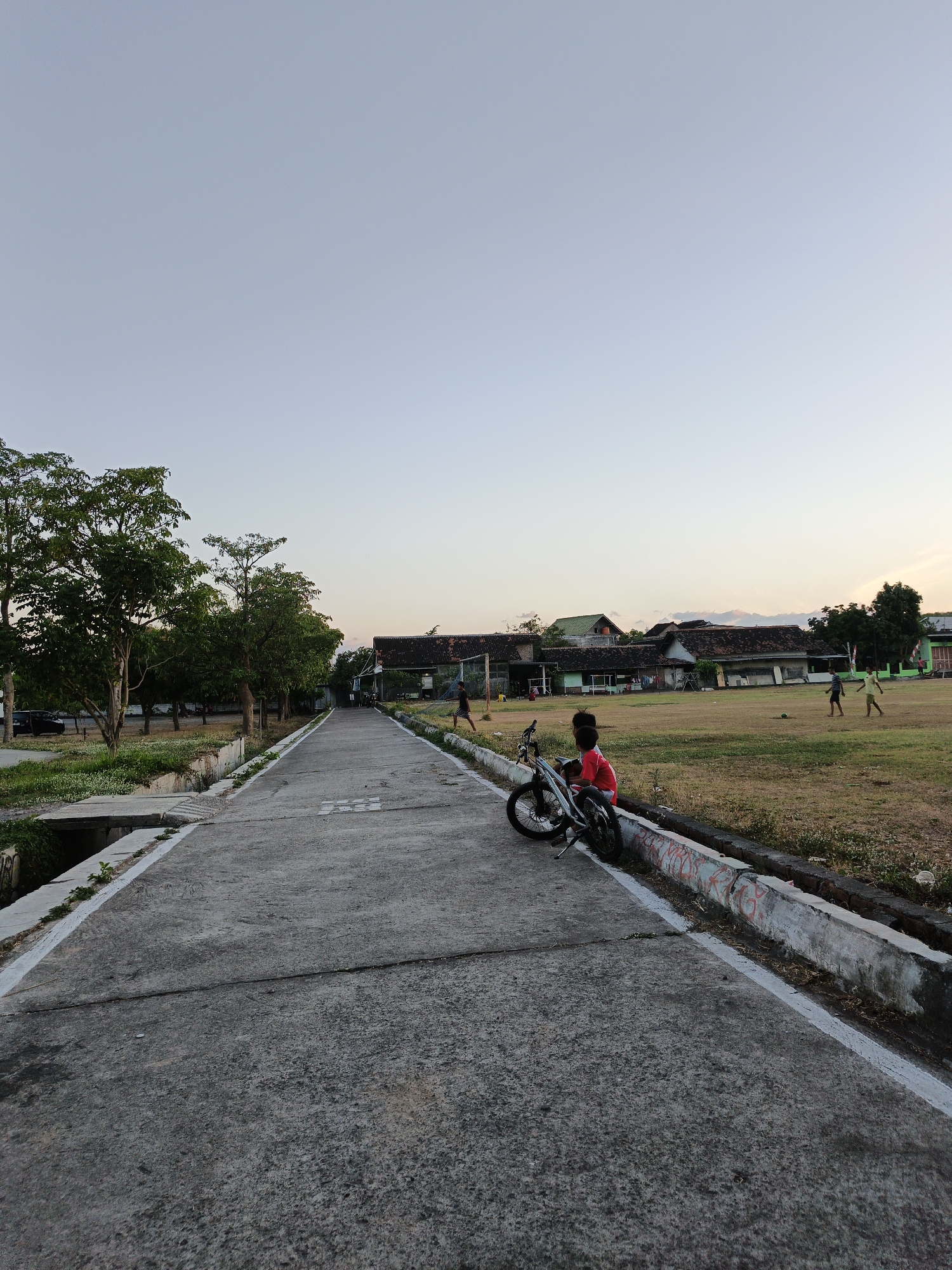 The image depicts a concrete road leading towards buildings and a grassy field. The sky is clear and light blue, suggesting it is either early morning or late afternoon. On the left side of the road, there are trees and some vegetation. On the right side, there is a grassy field where several children are playing. In the foreground, on the right side of the road, a young boy wearing a red shirt is sitting next to a bicycle. The buildings in the background appear to be residential or educational facilities with tiled roofs. The overall scene suggests a peaceful, everyday moment in a community setting in Klaten, Indonesia.