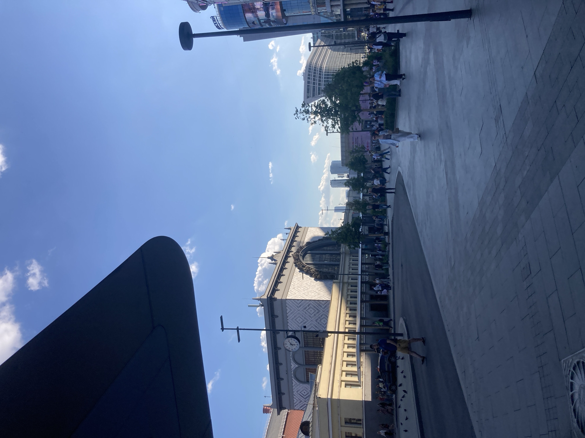 This image depicts a sunny urban scene in Moscow, Russia, specifically focusing on the area around Kazan Railway Station.
In the foreground, a dark, curved architectural element, possibly an awning or building overhang, occupies the upper-left quadrant. Below this, a grey tiled pavement covers the bottom-right portion, featuring a circular metal grate in the lower-left. A dark-colored asphalt road curves through the lower-left to mid-left, with a man in a blue t-shirt and light-colored shorts walking across it towards the right. Several pigeons are scattered on the road near him. A dark sedan is visible on the road behind the man.
The midground is dominated by the ornate facade of the Kazan Railway Station, a large light-colored building featuring a grand arched window with intricate details on its right side and geometric patterns across its upper walls. A round clock is mounted on a tall, black street light pole positioned directly in front of the station's left section. Numerous pedestrians, dressed in casual clothing, traverse the wide plaza and sidewalks in front of the station. A woman in a light blue suit and sunglasses walks towards the viewer on the grey pavement in the mid-right. Several trees with green foliage are planted in designated areas within the plaza.
In the background, to the right, a modern, multi-story building with a curved glass facade and large digital screens showing text like "BCKИЙ" and "СБЕРБАНК" rises prominently. Further in the distance, a cluster of tall, modern skyscrapers is visible against the horizon. The sky is clear and bright blue with scattered white cumulus clouds. The lighting indicates a bright day, casting long shadows from people and structures towards the right.