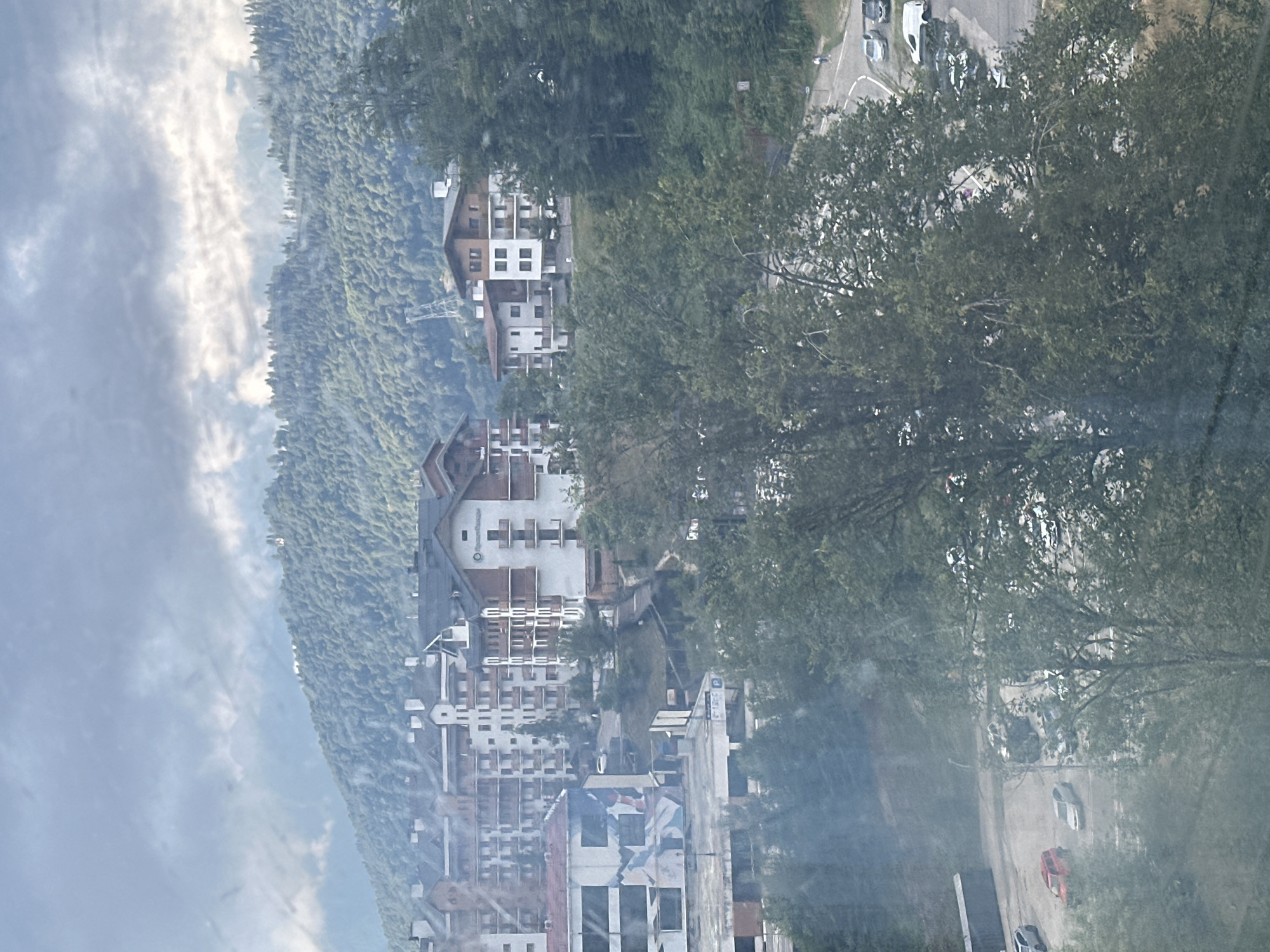 The image depicts a panoramic view of a mountainside resort town, likely Estosadok, Russia, characterized by a cluster of multi-story buildings with modern architecture nestled amidst lush green trees and rolling hills. The buildings, with their distinctive dark roofs and white or light-colored facades, appear to be residential or hotel complexes, some featuring balconies and large windows. The natural environment is dominant, with dense forests covering the slopes of the mountains that rise in the background under a cloudy sky. In the foreground and mid-ground, trees create a natural screen, partially obscuring the view of the buildings and a road that winds through the landscape. Several cars are visible in a parking lot near one of the buildings, and a few individuals can be seen walking on a path. The overall impression is one of a well-developed mountain resort, blending modern construction with a scenic natural setting.