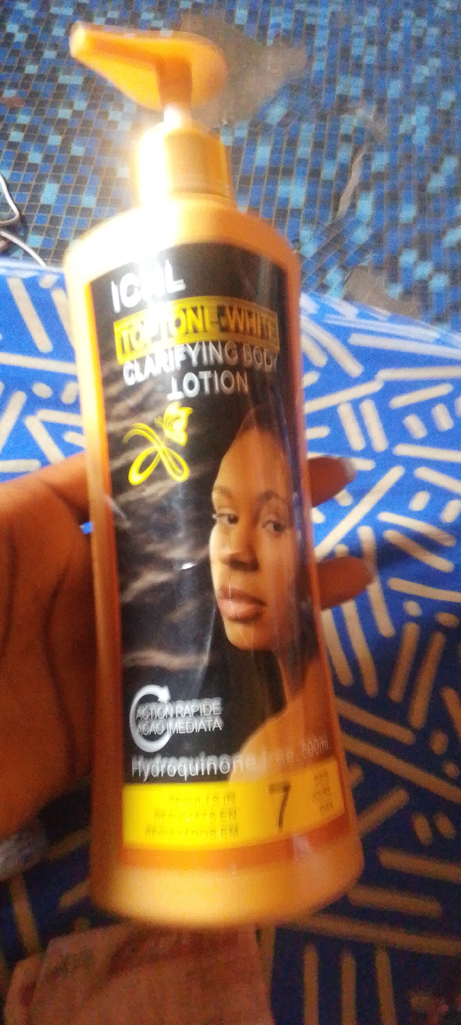 The image shows a person holding a bottle of "ICAL TOPTONE WHITE CLARIFYING BODY LOTION". The bottle is orange and has a black label with white and yellow text. The label features the brand name "ICAL", the product name "TOPTONE WHITE CLARIFYING BODY LOTION", and an image of a woman's face. Below the image of the woman, there is text that reads "ACTION RAPIDEACAO IMEDIATA" and "Hydroquinone free 500ml". There is also a graphic with the number "7" and the word "DAYS" next to it. The background appears to be a blue and black tiled surface. The lighting suggests it could be daytime, but the specific time and location are not discernible. The overall impression is a product shot, likely for advertising or personal use.