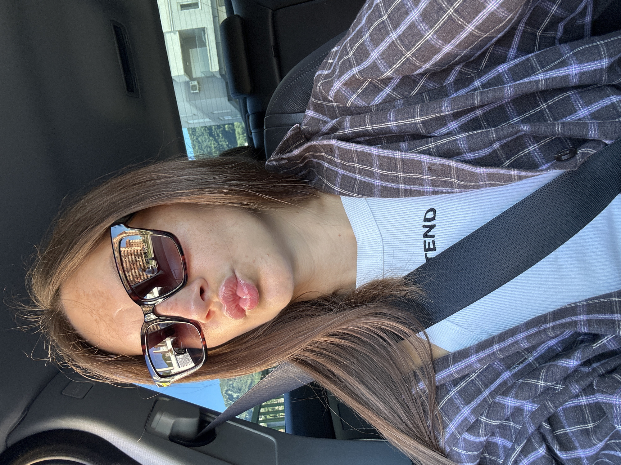 A young woman with brown hair is sitting in the passenger seat of a car, wearing sunglasses, a white ribbed top with the word "TEND" printed on it, and an open plaid shirt. She is puckering her lips and sticking out her tongue slightly. The interior of the car is visible, including a seatbelt and part of the dashboard. Outside the car, a residential building and a tree-covered hillside can be seen, suggesting a location in a town or city, possibly Donji Pobori, Montenegro. The scene appears to be captured during daylight.