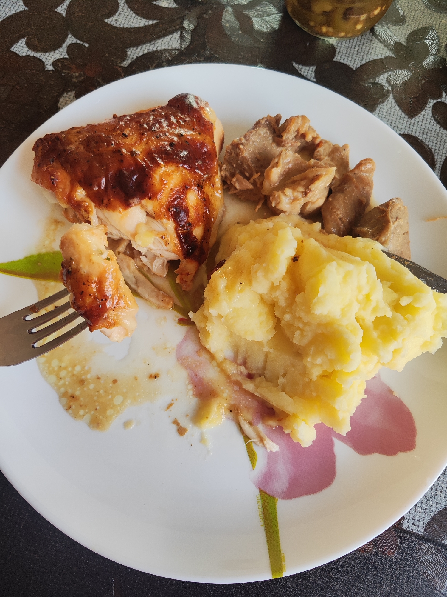 A close-up, eye-level shot captures a white plate filled with a meal. On the left is a piece of golden-brown roasted chicken, its skin glistening, with a fork holding a small piece of chicken to the side. To the right of the chicken are several pieces of cooked meat, likely stewed or braised, with a light brown color. In the foreground and center of the plate is a generous serving of mashed potatoes, a pale yellow color, with visible texture from mashing. The plate has a floral design of a pink and purple flower with a green stem. The background shows a dark patterned placemat and a sliver of a glass jar with green contents. The lighting is natural and appears to be from overhead. The scene is likely indoors, on a table, in Gvardeysk, Russia.