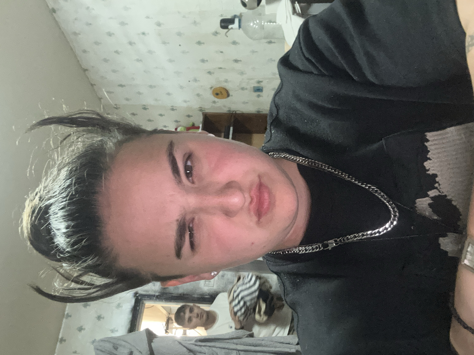 A person with short, slicked-back dark hair, wearing a black t-shirt and a silver chain necklace, is positioned in the foreground. Their right eye is squinted, creating a winking expression. Behind them, visible in a mirror, is another person wearing a white shirt. The background features patterned wallpaper, and objects on shelves. The location is not identifiable as Plovdiv, Bulgaria from the image.