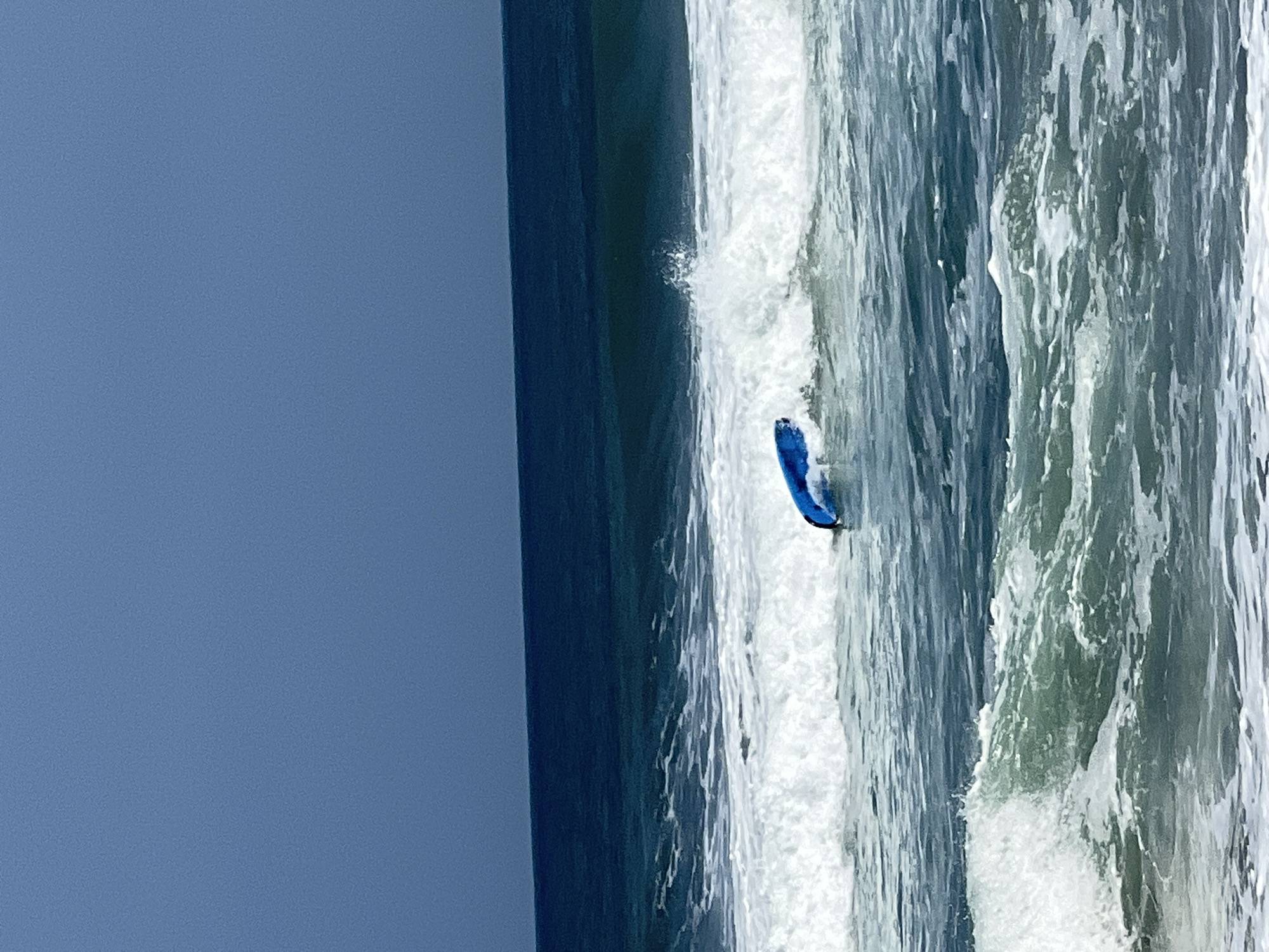A blue surfboard is partially submerged in the ocean waves off the coast of Kuta, Indonesia. The sky is a deep, overcast blue, and the ocean is a dark, turbulent teal. White foam from the breaking waves surrounds the surfboard, which appears to be riding a wave or possibly capsized. The scene depicts a dynamic ocean environment with a single, brightly colored object amidst the natural elements.